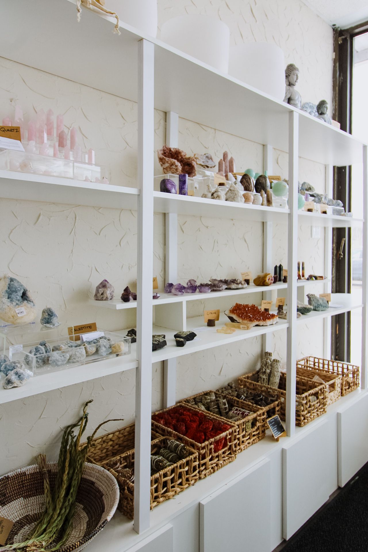 White retail shelving unit displays crystals, stones, glassware and decorative items with woven baskets on bottom shelves.