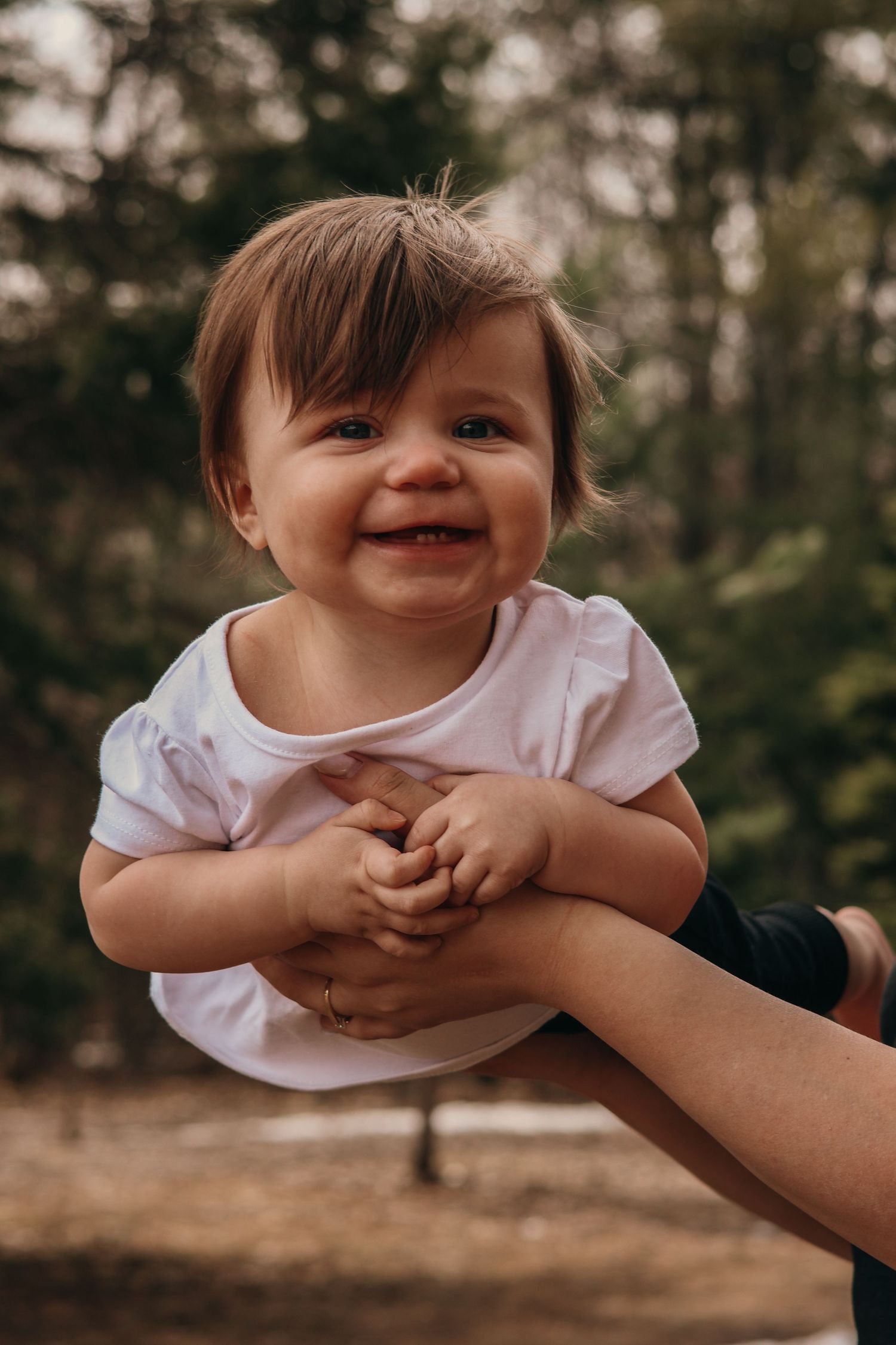 Sweet baby in a white shirt smiles joyfully while being held up outdoors against a blurred forest background.