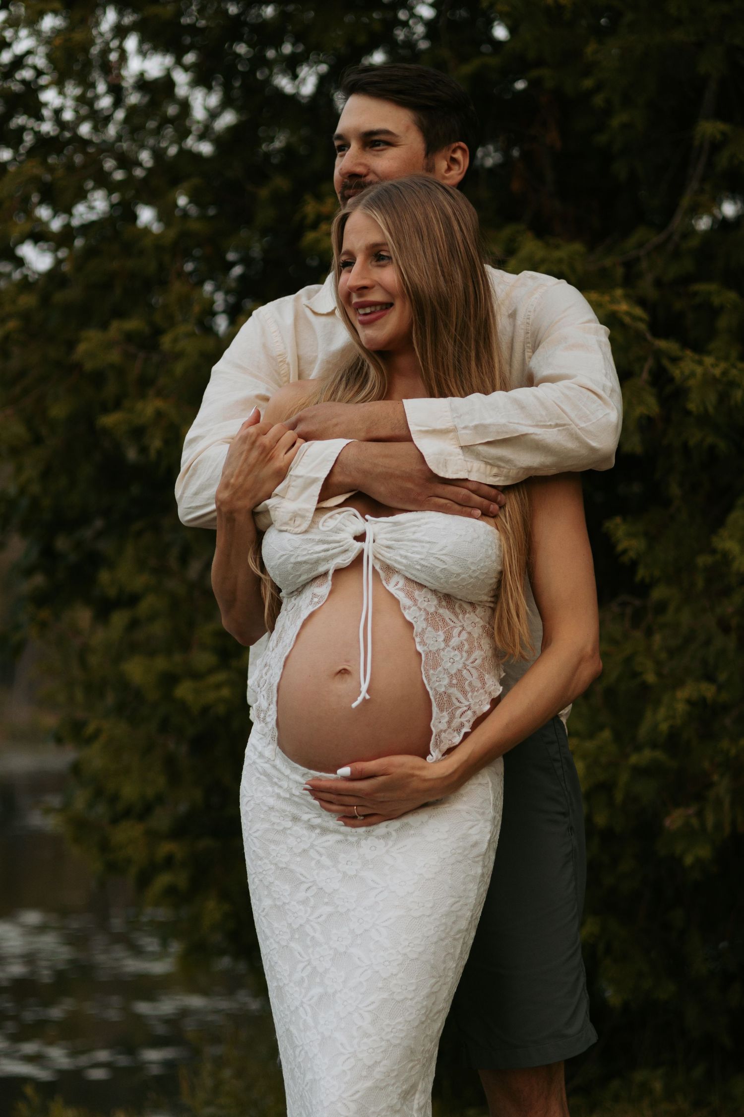 Romantic maternity photo outdoors with couple embracing in white lace dress by tranquil water at sunset.