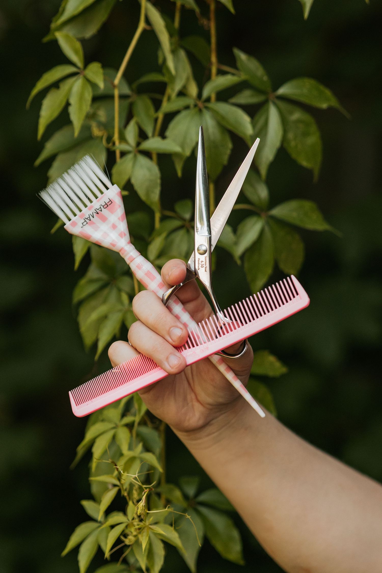 Hands holding white and pink plastic barber scissors and combs against green foliage background.