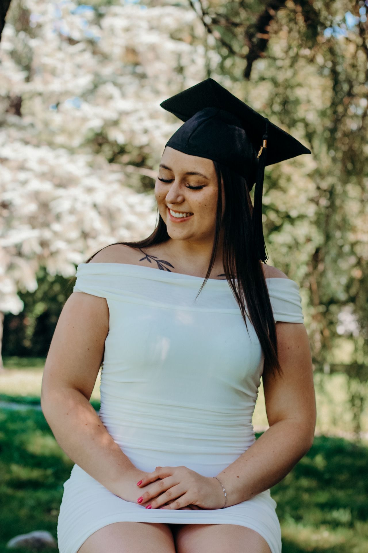 Graduate in white off-shoulder dress and black cap smiling outdoors in front of blooming spring trees on sunny day.