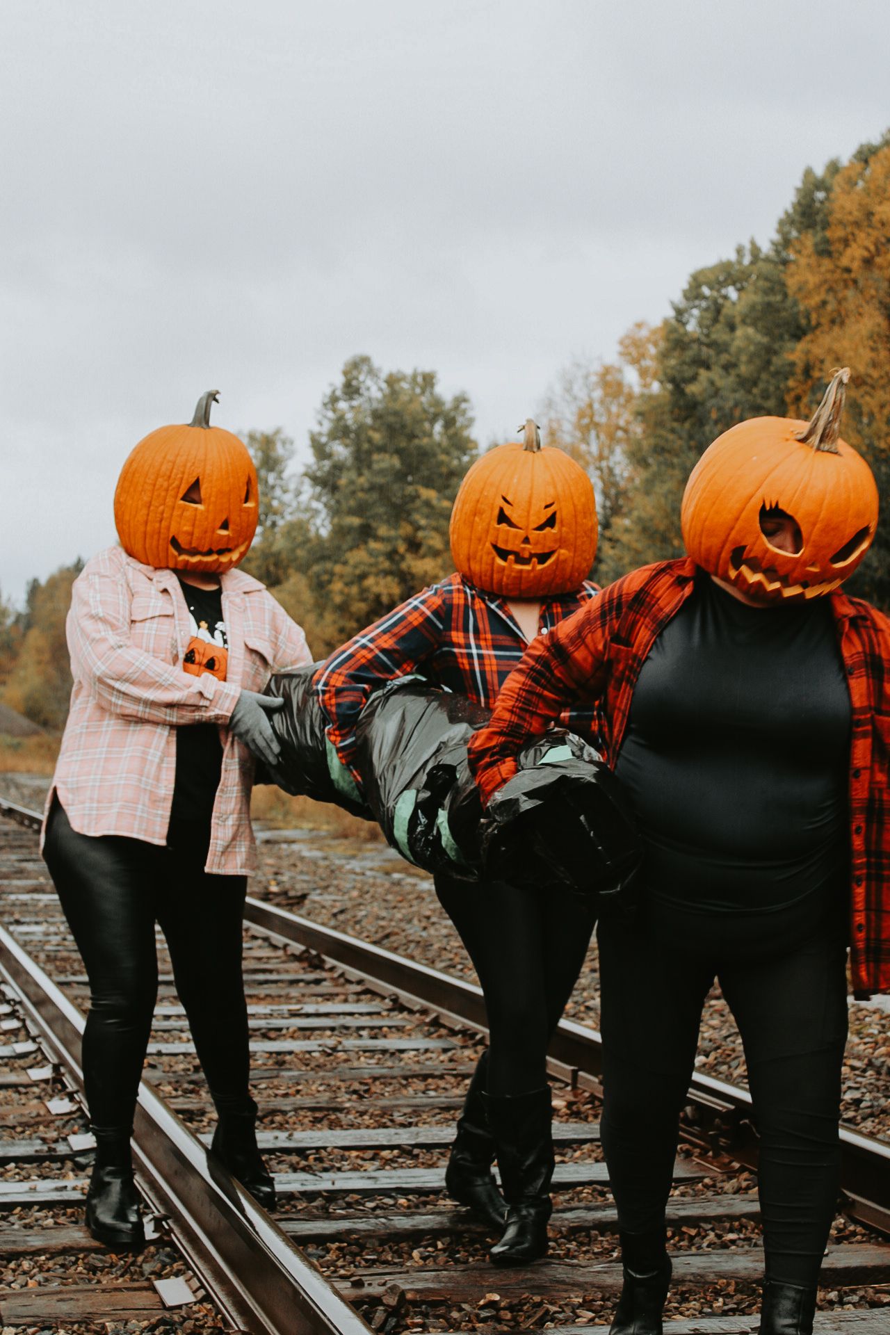 Three people wearing pumpkin heads and plaid shirts pose playfully on railroad tracks.