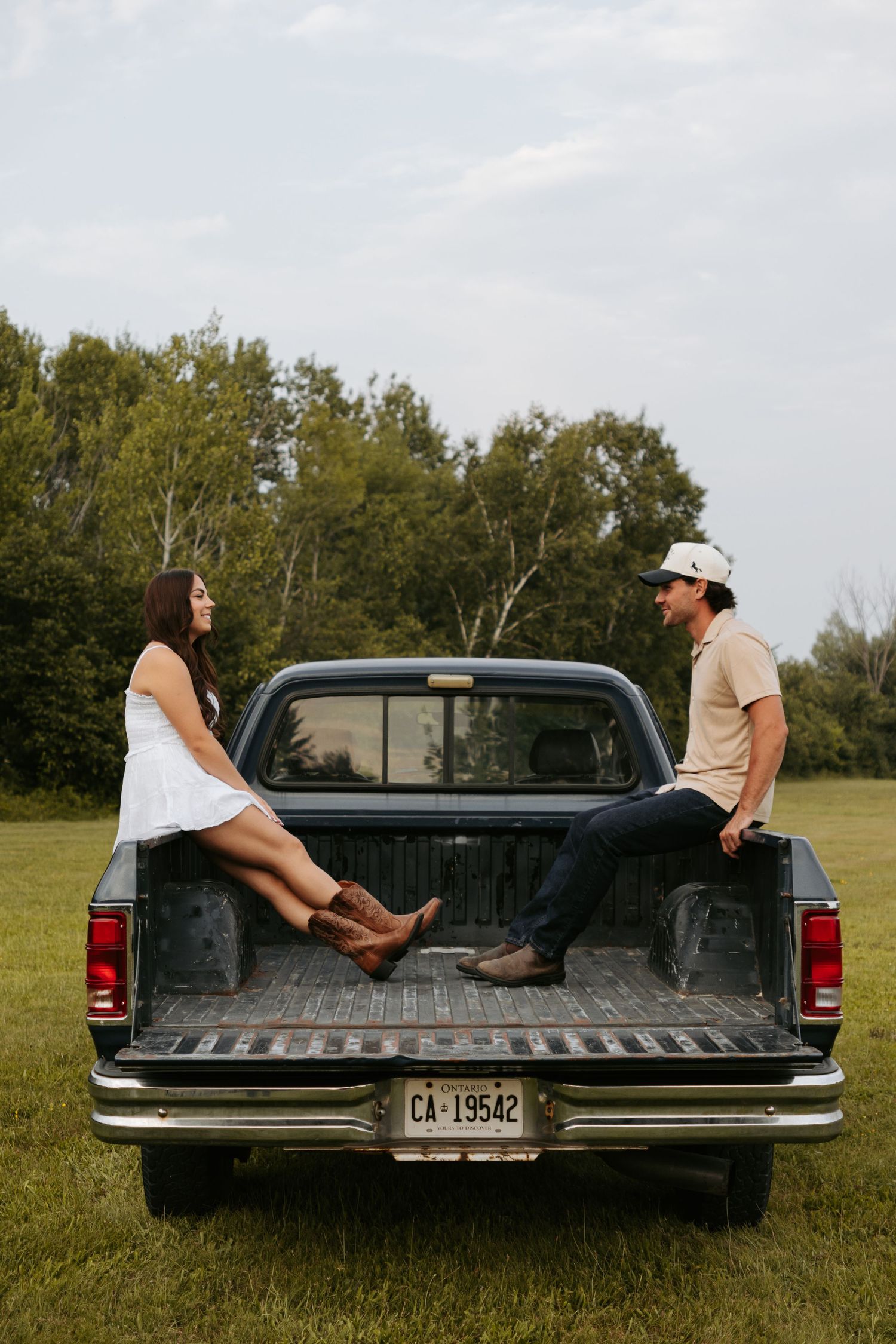 Two people sit in bed of vintage pickup truck in countryside field having casual conversation.