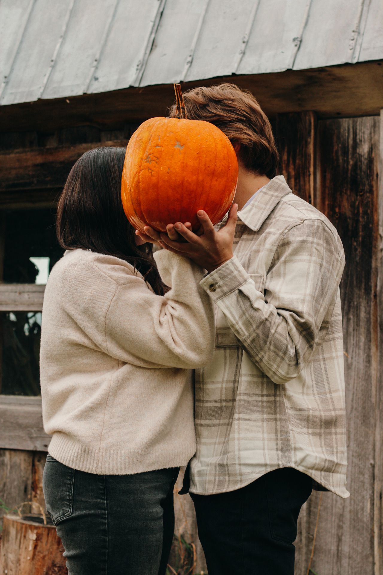 Couple hiding their faces behind a large orange pumpkin in front of a rustic wooden wall.