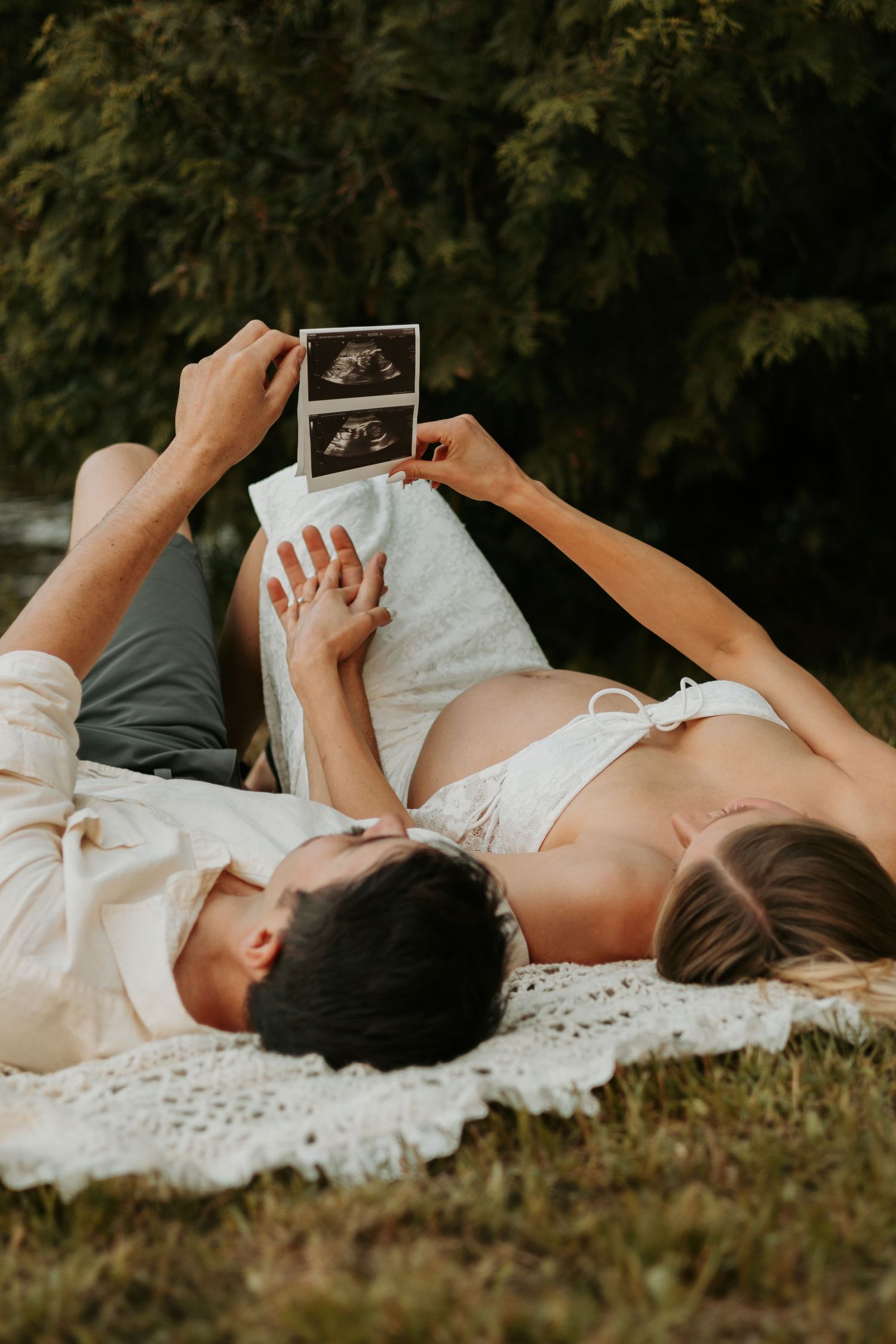 A couple lies on a blanket outdoors holding ultrasound photos while enjoying a tender pregnancy moment.