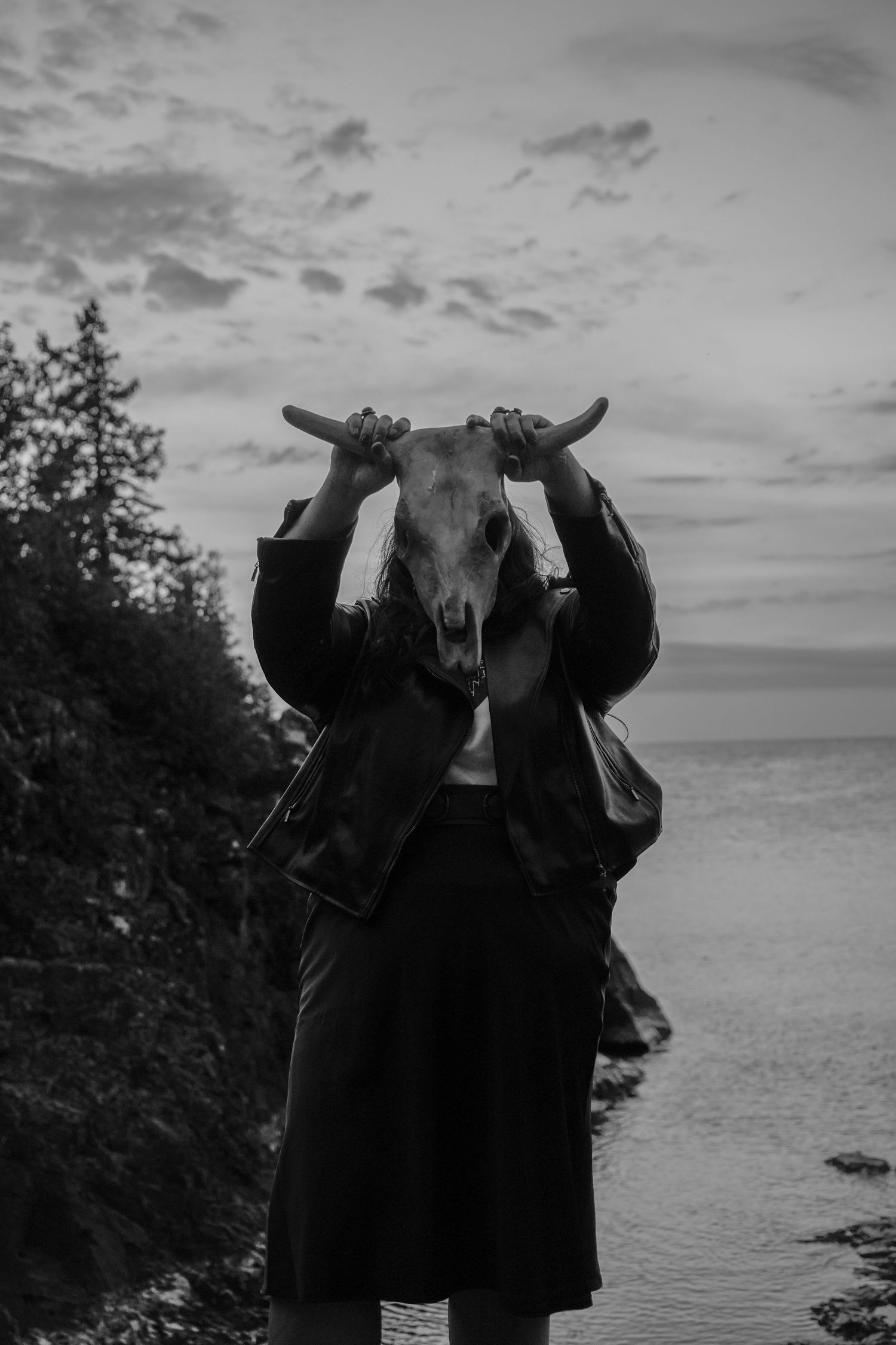 A dark figure wearing a long coat holds an animal skull against a moody coastal landscape at dusk.