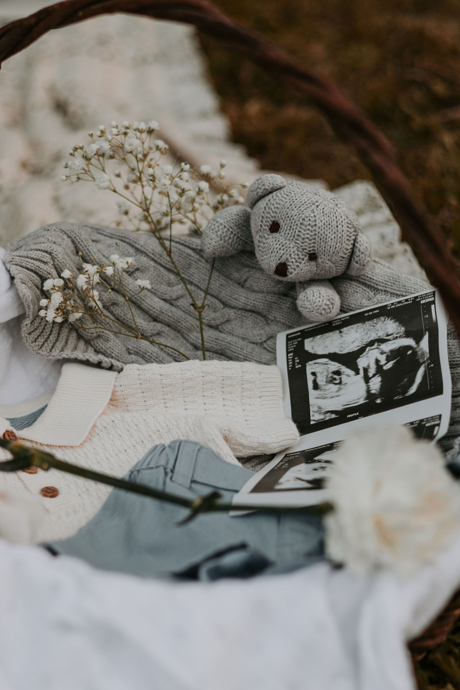 Baby announcement setup with grey knit teddy bear, dried flowers and ultrasound photos on white fabric.