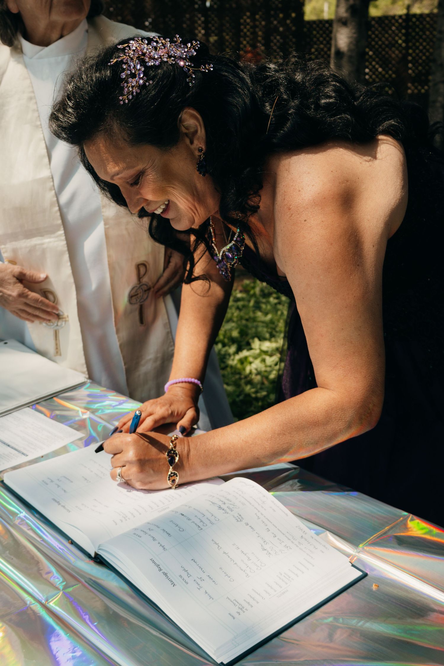 Someone in a black dress bends over a guest book to sign while smiling during an outdoor event.