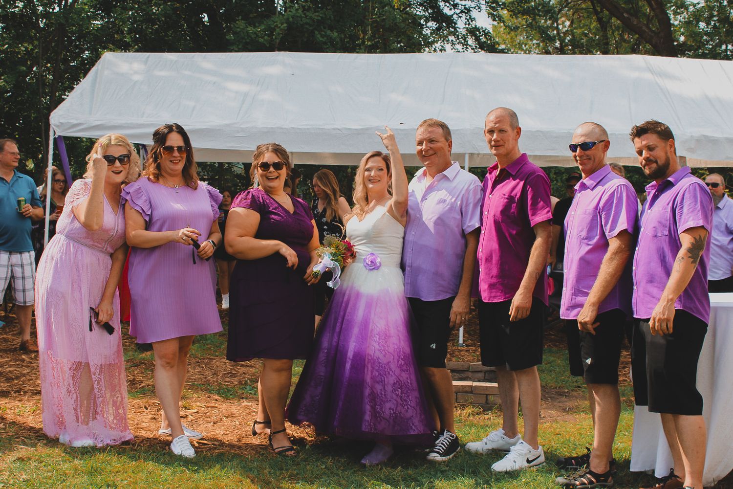 A bridal party wearing coordinating purple and pink outfits poses together outdoors at a casual summer wedding celebration.