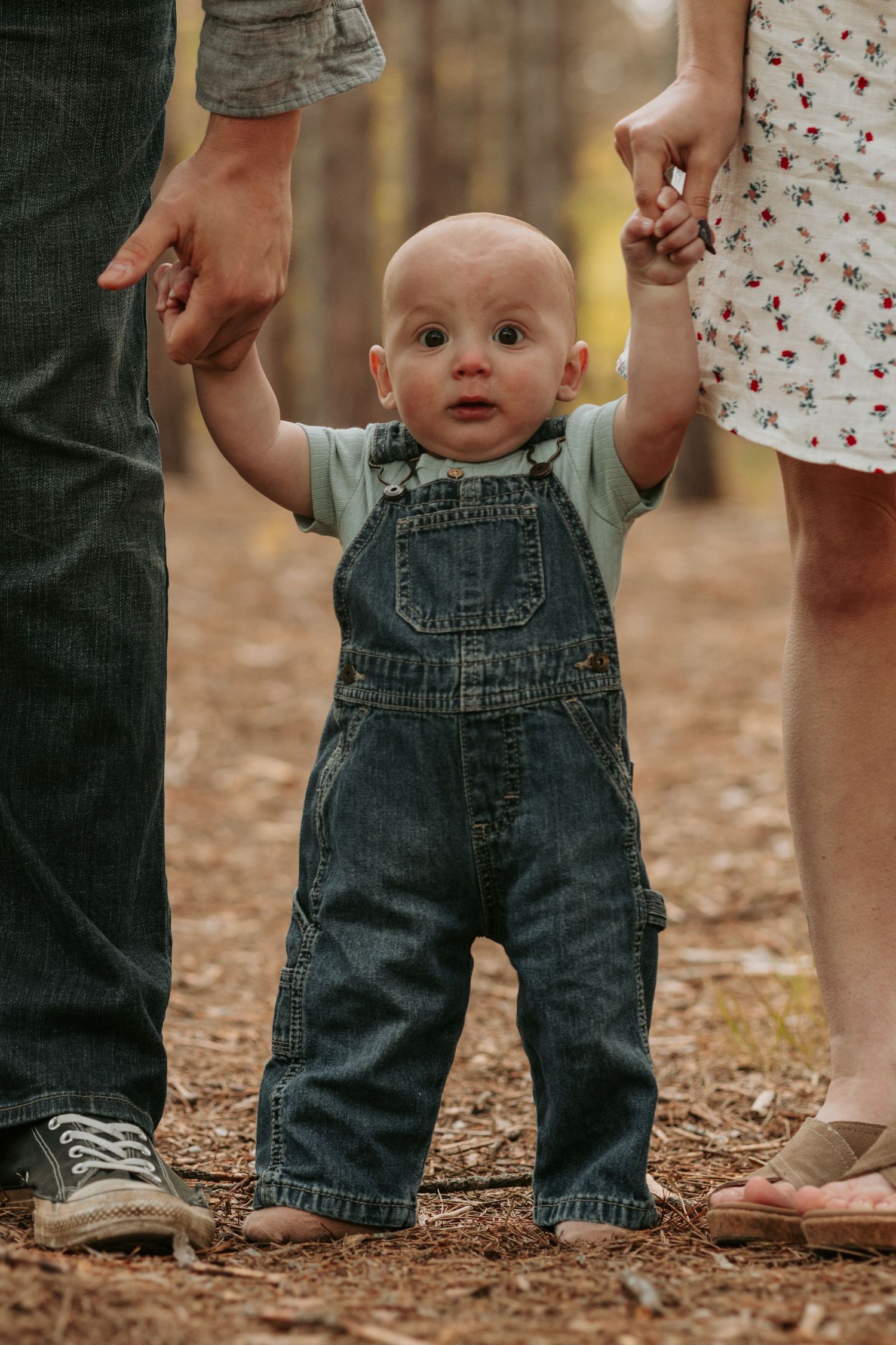 Small legs wearing denim overalls standing on autumn ground while being supported by adults.