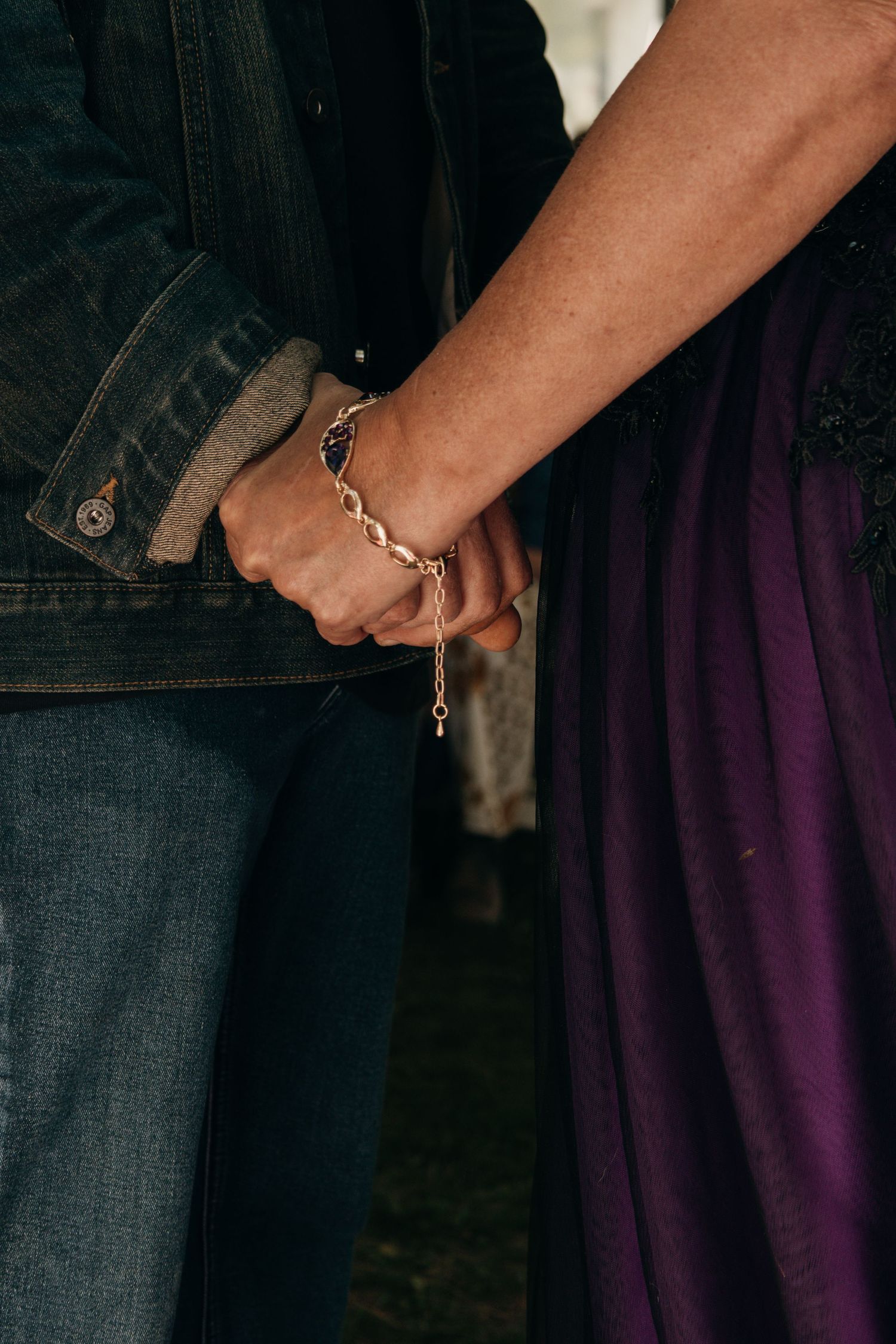 Close-up shot of two people holding hands, one wearing a denim jacket and the other a purple dress with silver bracelet.