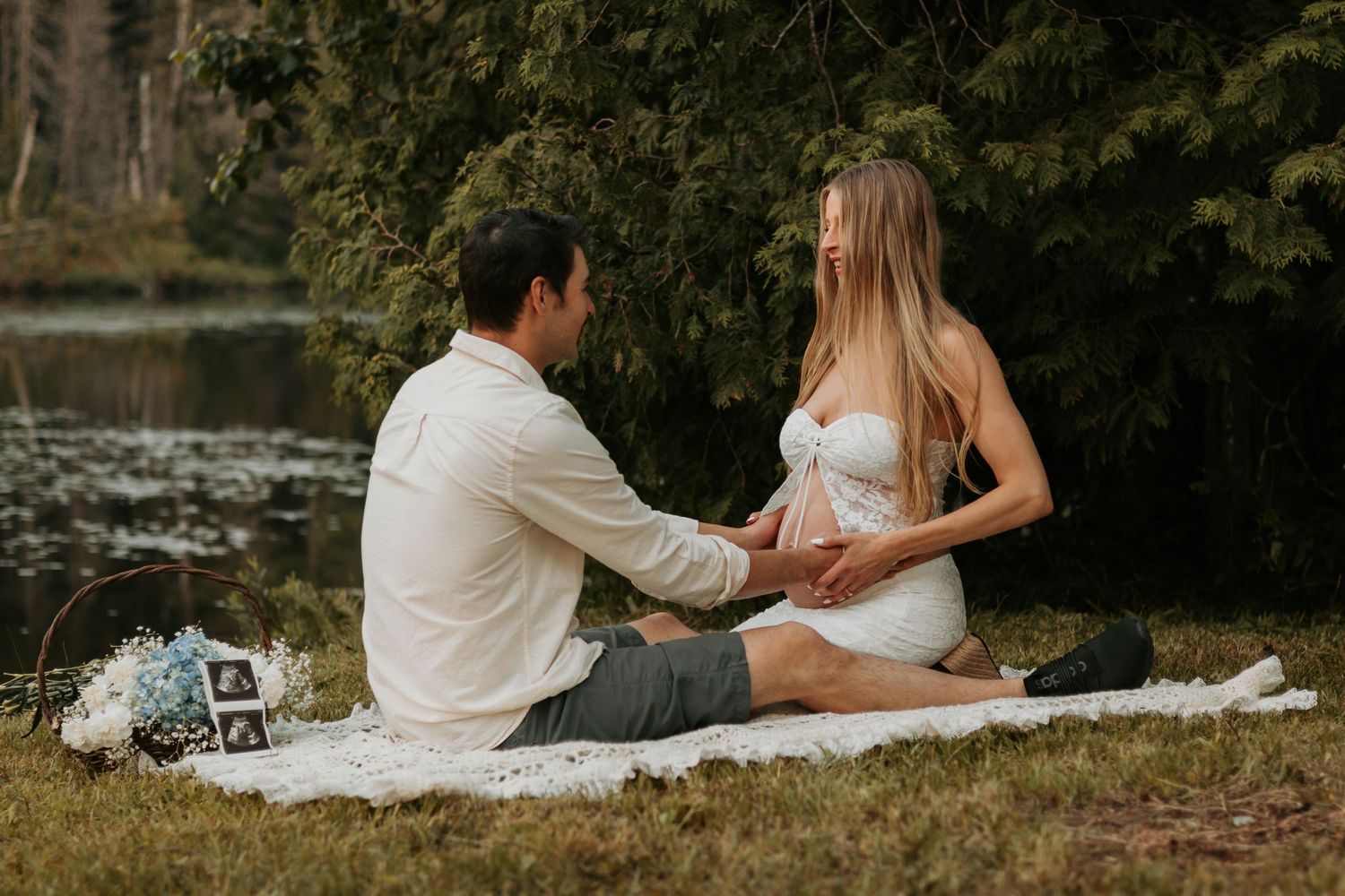 A couple sits together on a picnic blanket beside a serene lake surrounded by lush greenery on a warm day.