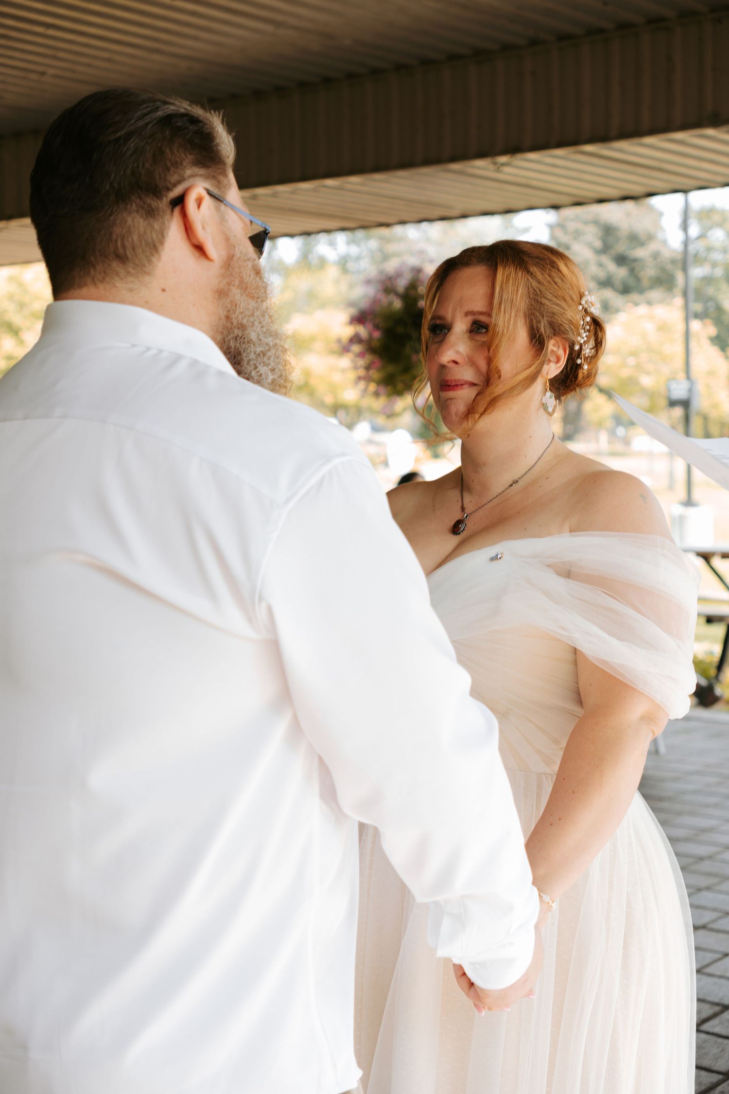 A romantic moment is captured as a couple in white attire embraces under an outdoor pavilion on their wedding day.