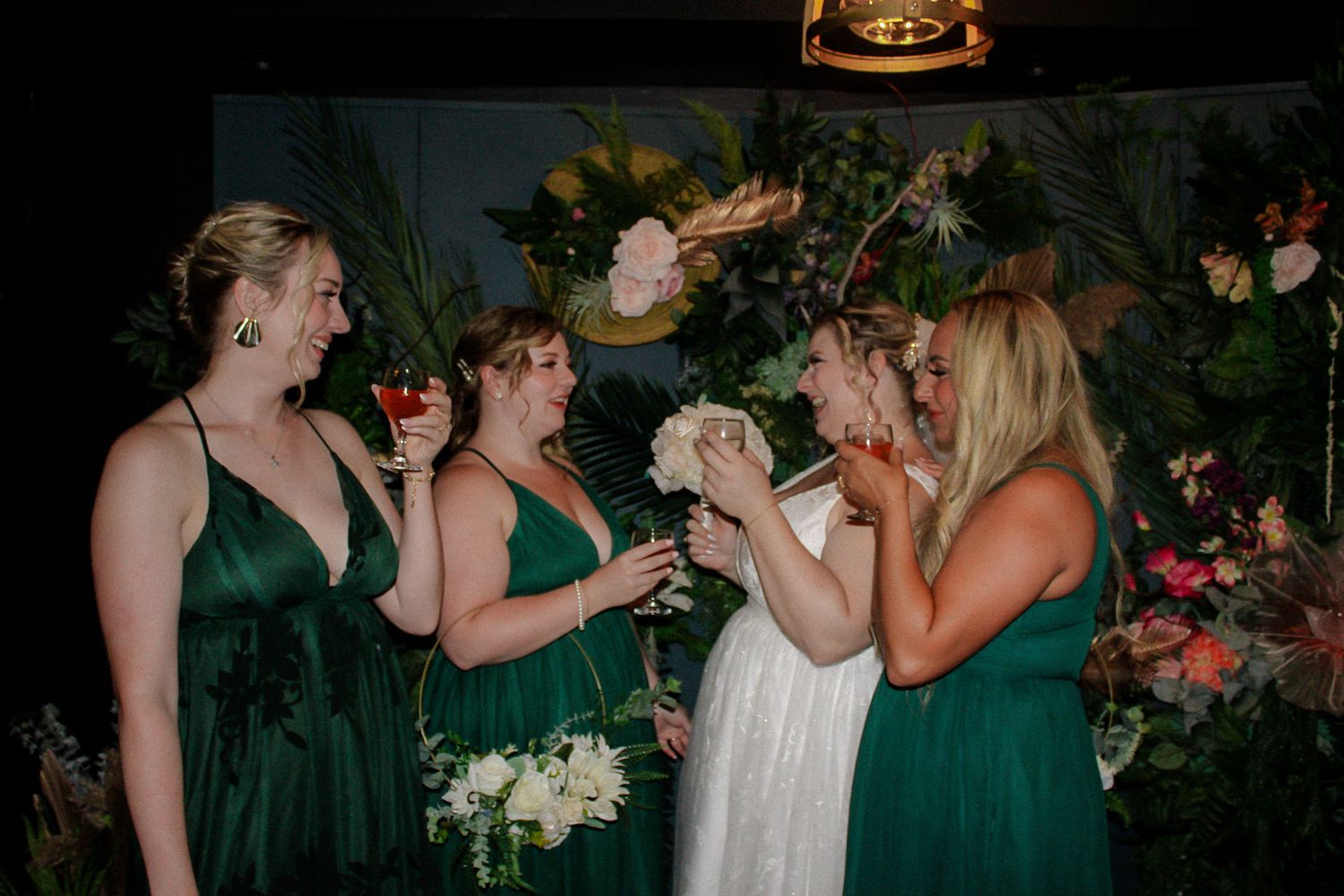 A group wearing emerald green dresses share a celebratory moment with drinks against a floral backdrop at an event.