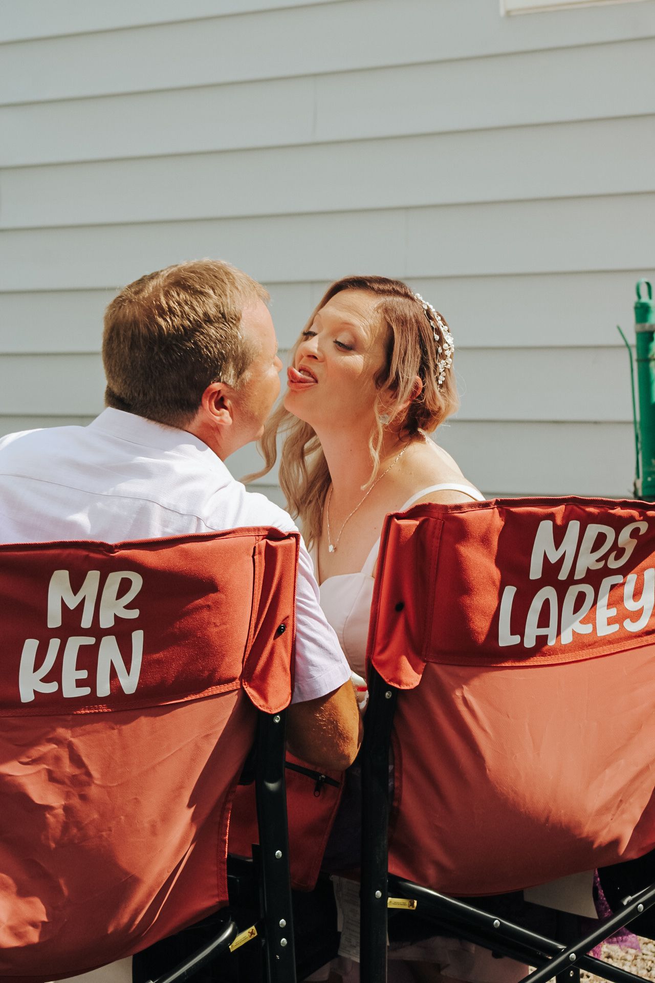 A couple shares an intimate moment while sitting in custom labeled directors chairs outdoors.