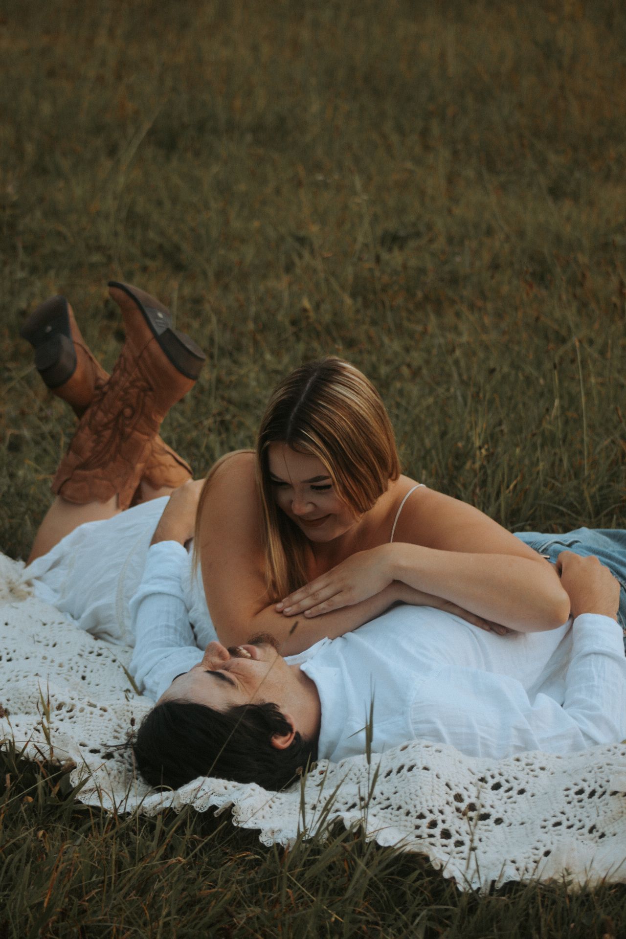 Romantic couple lying on white lace blanket in golden grass field.