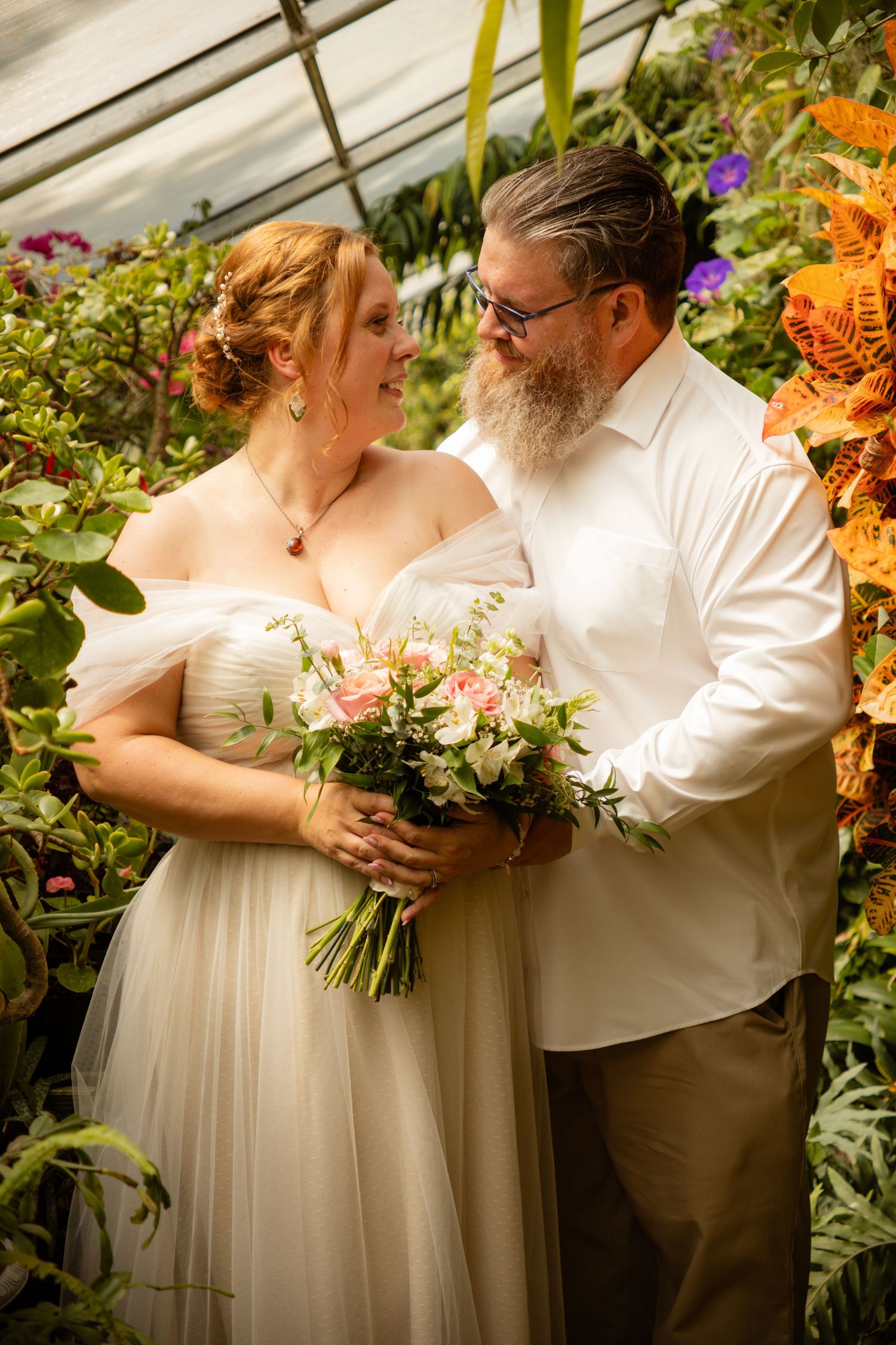 A couple shares an intimate moment in a greenhouse surrounded by colorful flowers as they hold a pink floral bouquet.