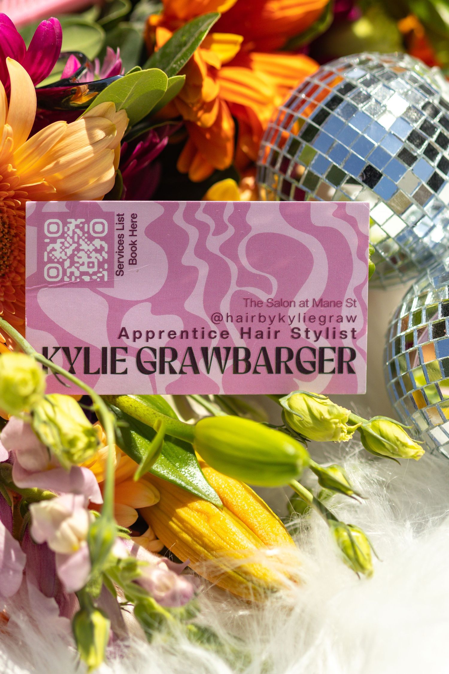 Pink business card for apprentice hair stylist displayed with fresh flowers and disco ball mirror decoration.