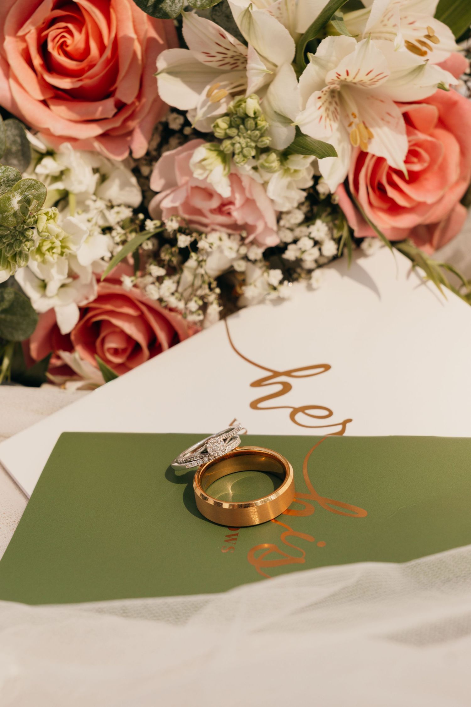 Wedding rings on green stationery with coral roses and white alstroemeria flowers in elegant bouquet arrangement.