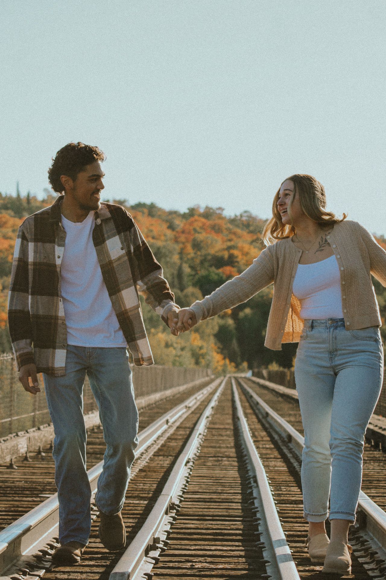 Couple walking hand in hand on railroad tracks with fall foliage in background.
