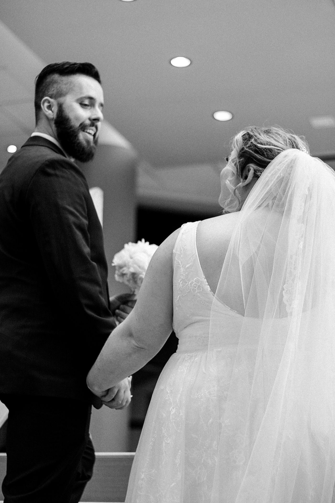A black and white photo showing a person in a white wedding dress and veil being helped with final adjustments.