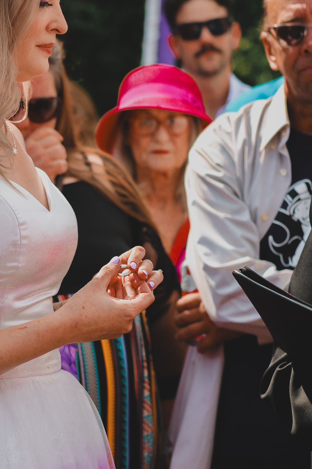 Woman in white dress holds small jewelry or ring at outdoor gathering with attendees in bright colors and sunglasses.