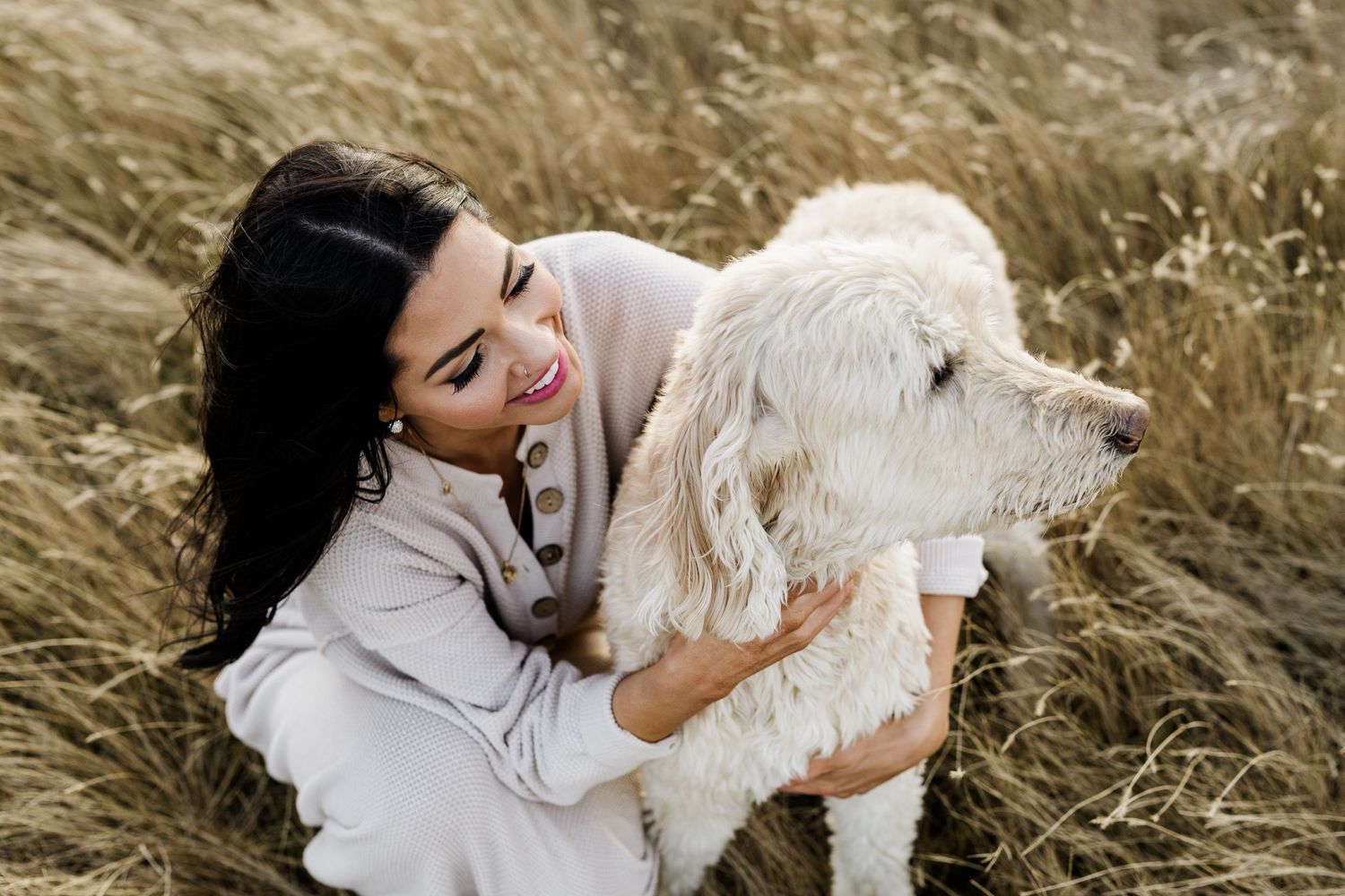Woman in light colored dress embracing white dog in golden field at sunset.