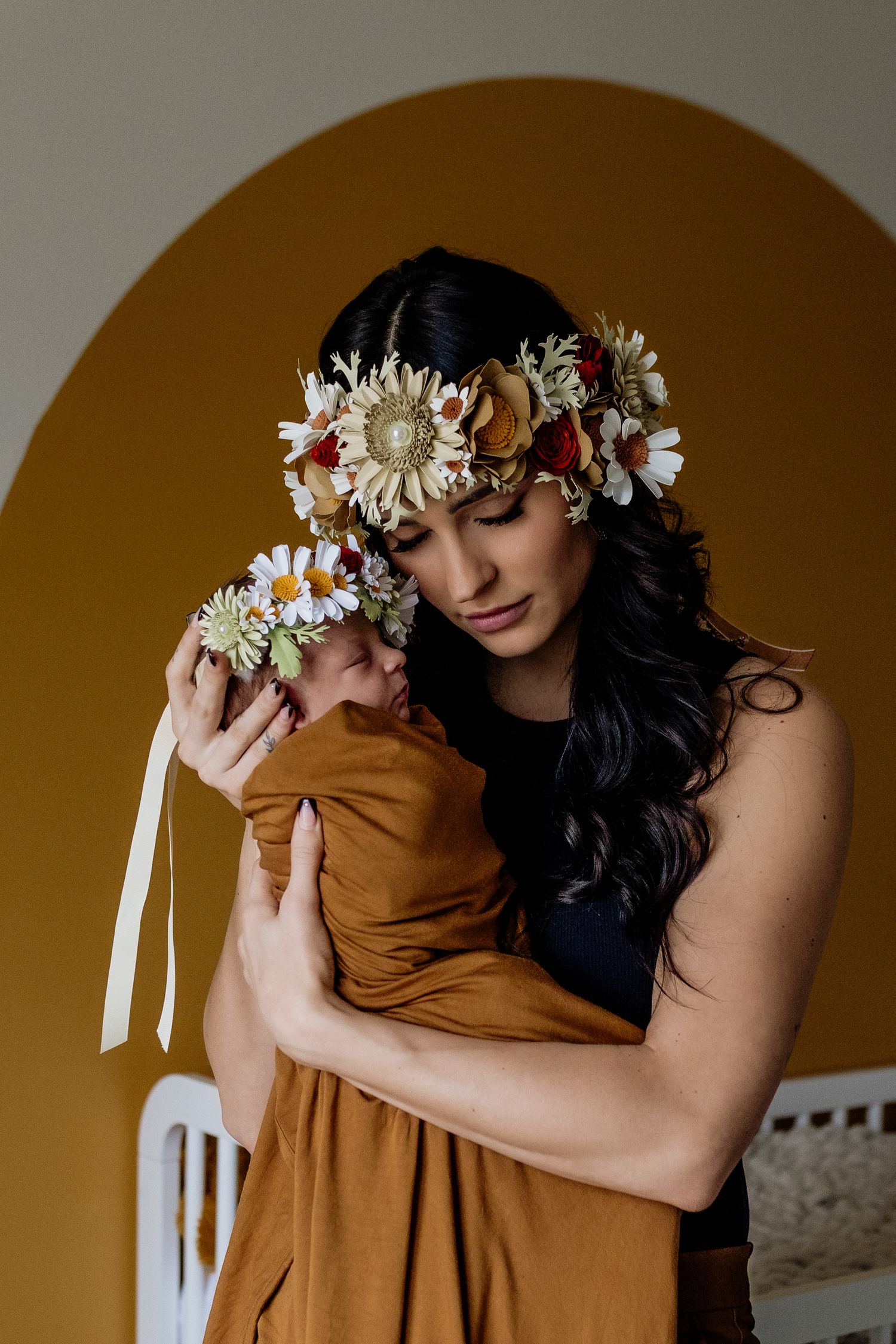 Woman wearing floral crown holds baby wrapped in mustard yellow fabric against warm brown background.
