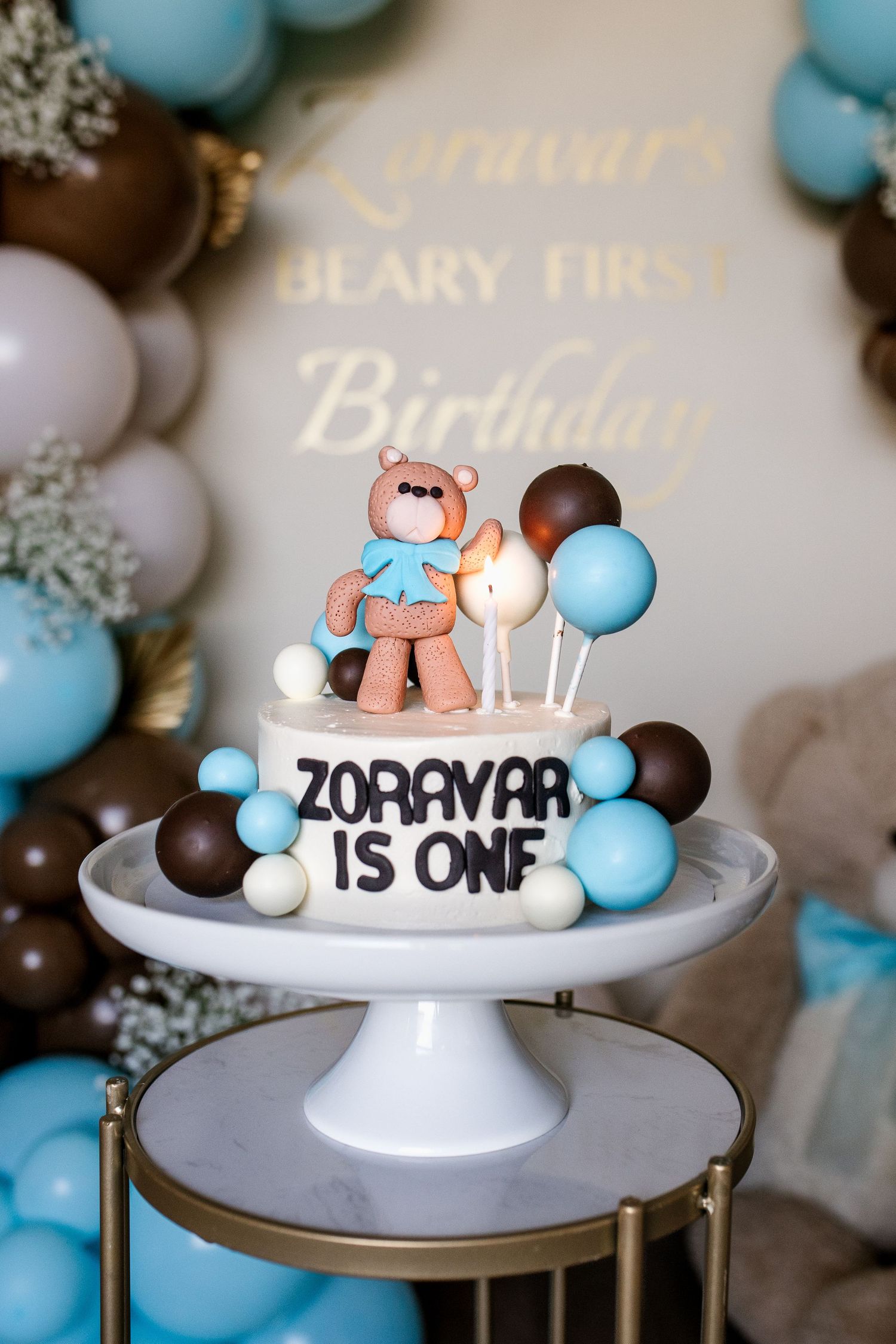 White cake stand displays a birthday cake decorated with blue and white balloons and teddy bear topper for first birthday.