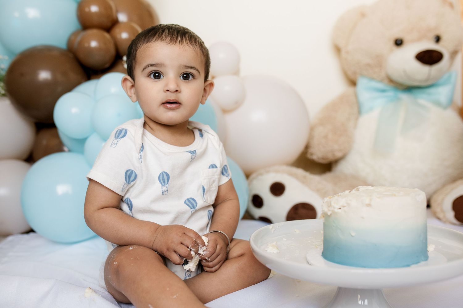 Baby in white onesie sitting among teddy bears and blue balloons.