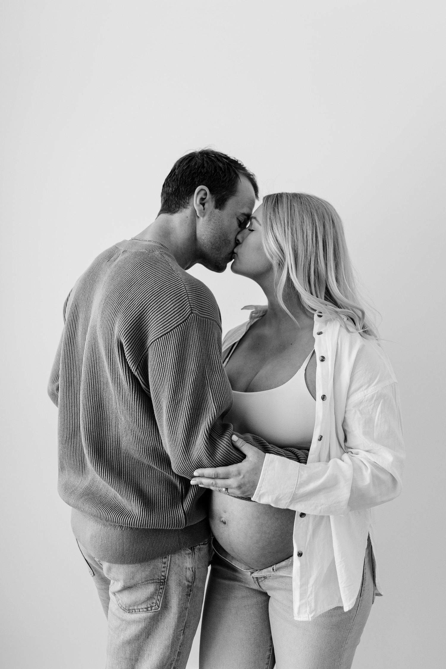 An intimate maternity photoshoot featuring a couple in casual attire against a white backdrop.
