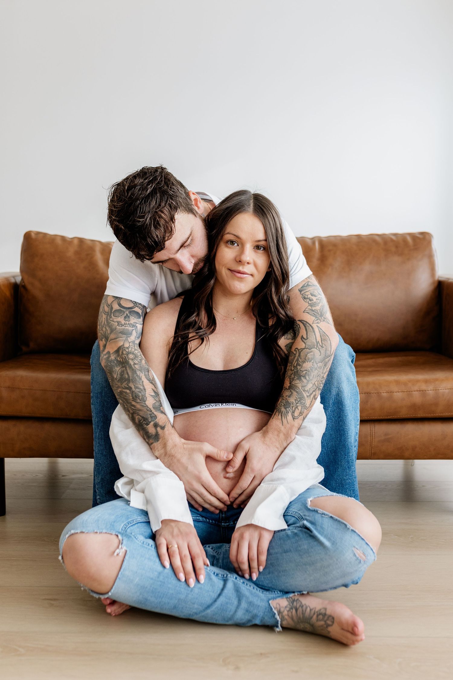 Couple sitting together on hardwood floor in front of brown leather couch in casual attire.
