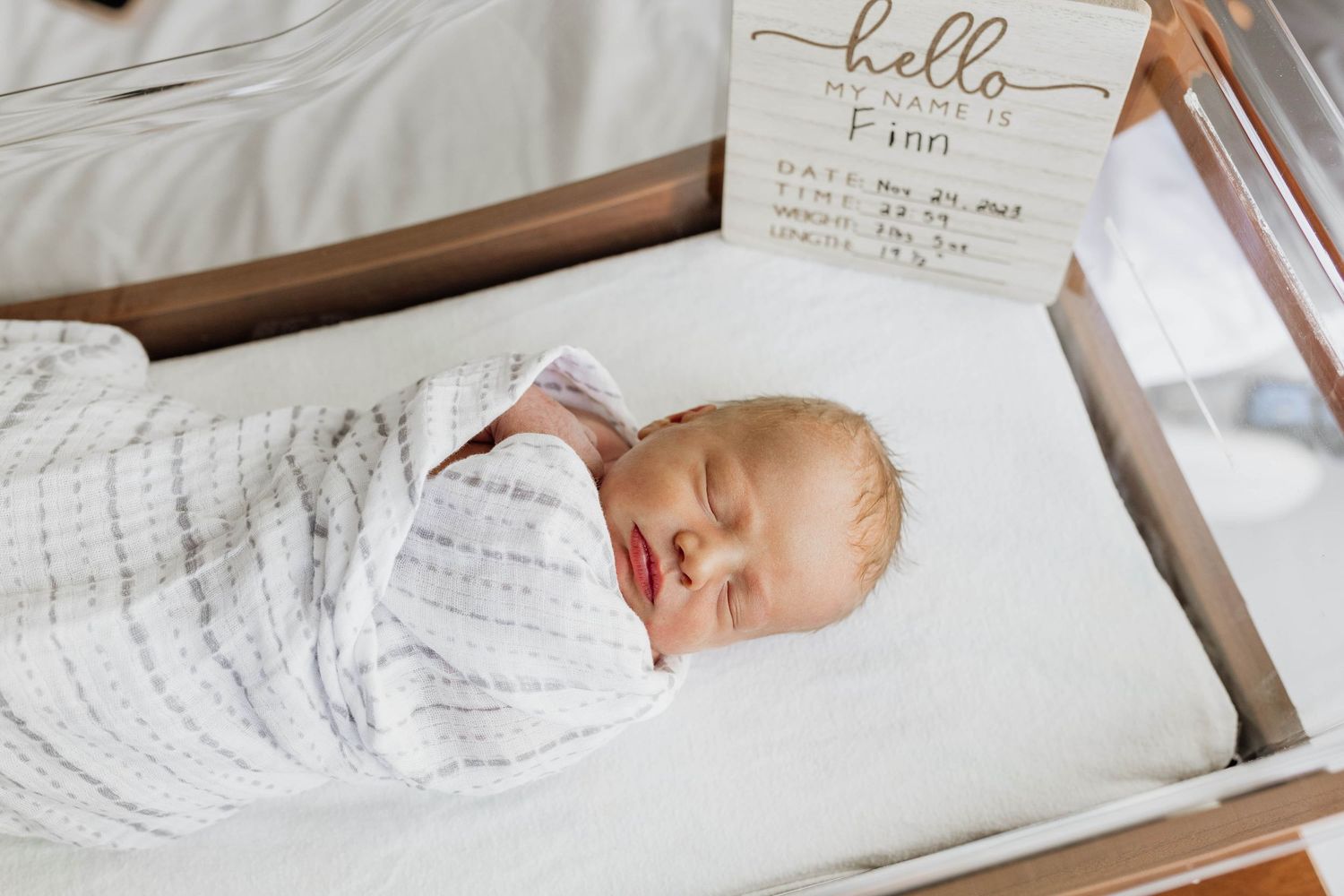 Newborn baby sleeping peacefully in a white bassinet with handwritten welcome sign.