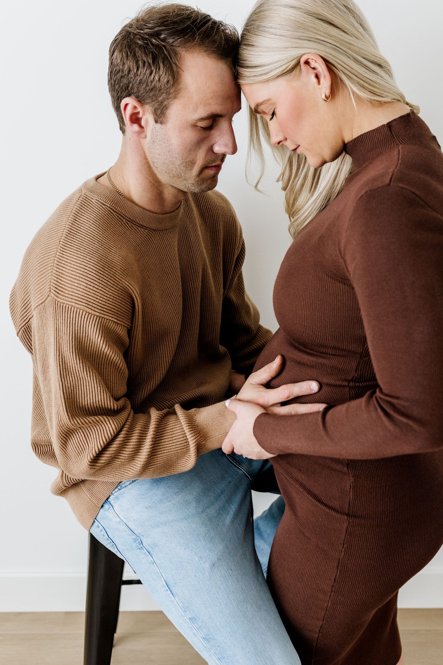 A couple in brown and tan sweaters share an intimate moment in a minimalist setting against a white wall.