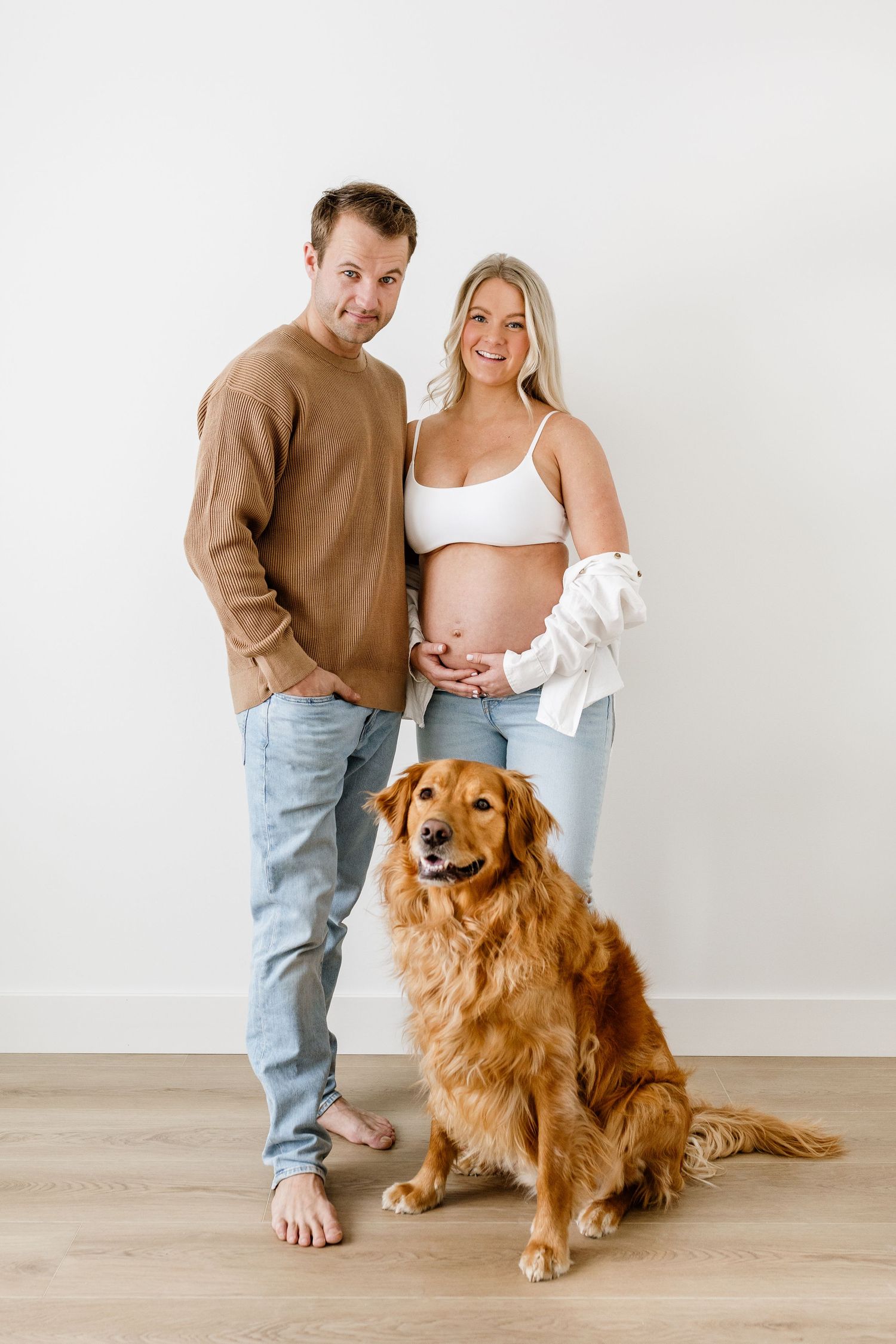 A couple poses with their golden retriever dog in a minimalist white studio setting for maternity photos.