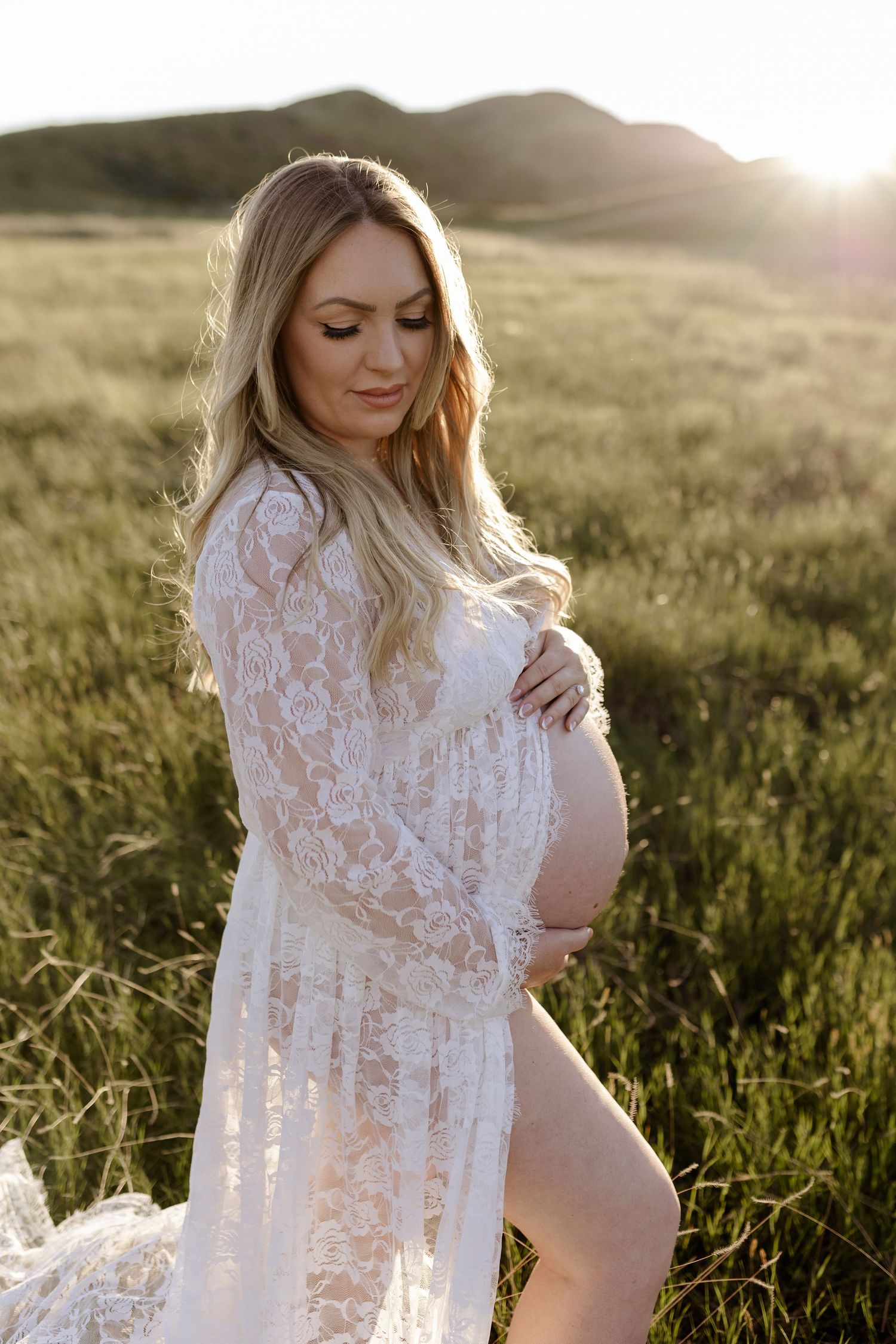 Maternity portrait in a white lace dress amid golden sunset light in a grassy field.