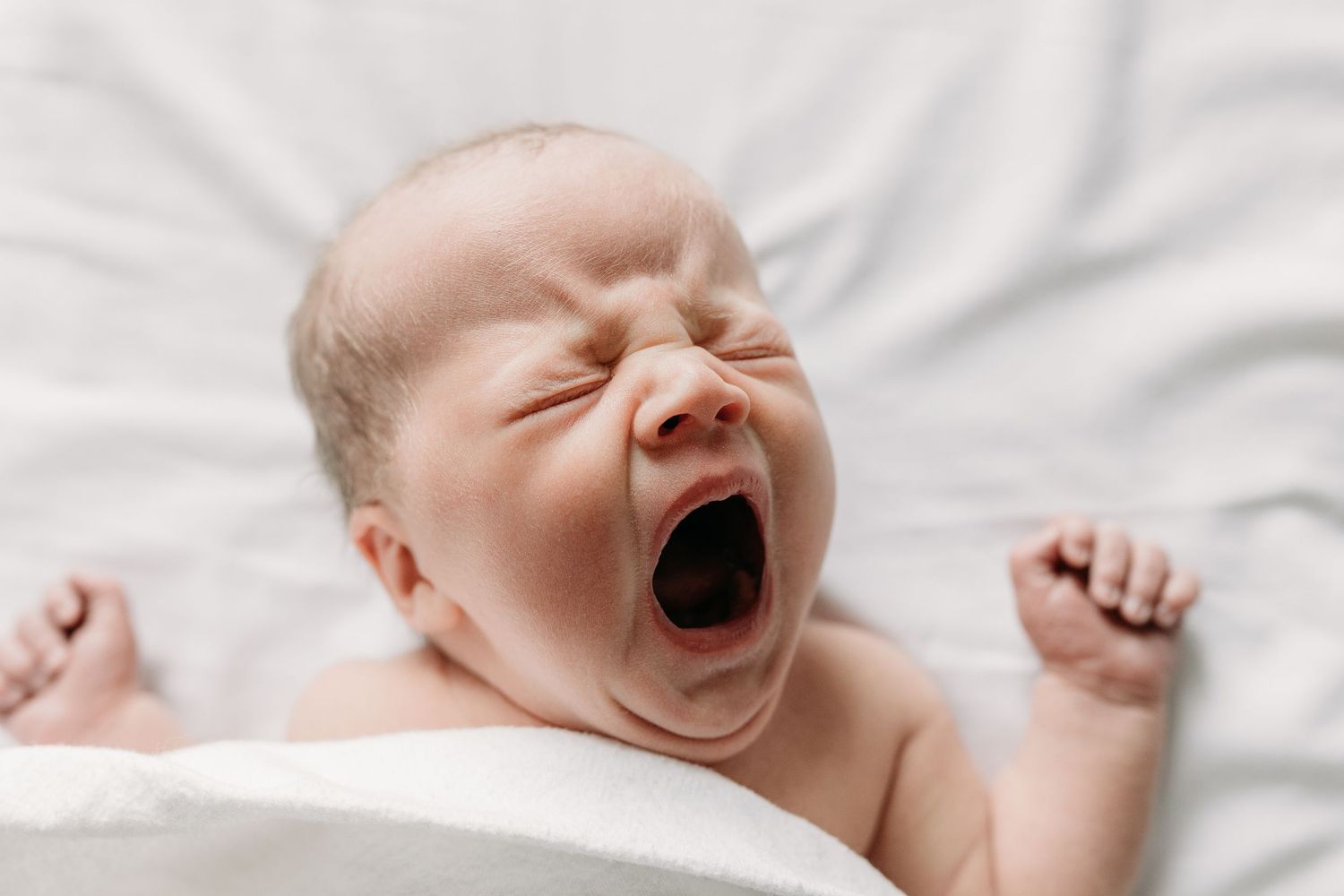 Newborn baby lying on white bedding with a wide yawn and stretched arms.