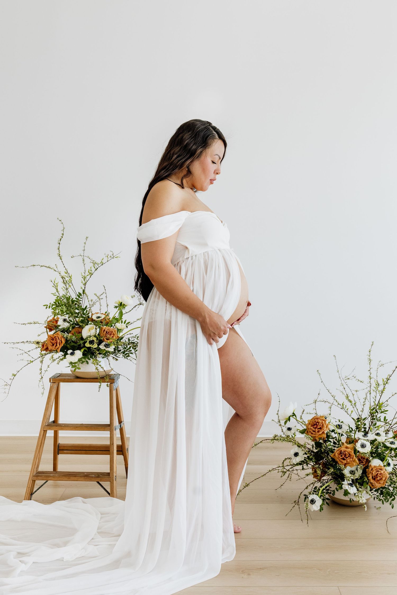Artistic maternity photos with white flowing fabric and floral arrangements on wooden stool.