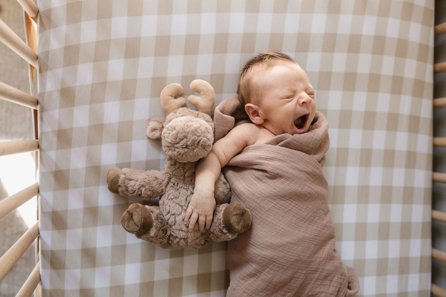 Sleeping baby cuddling with teddy bear on checkered blanket in soft natural light.