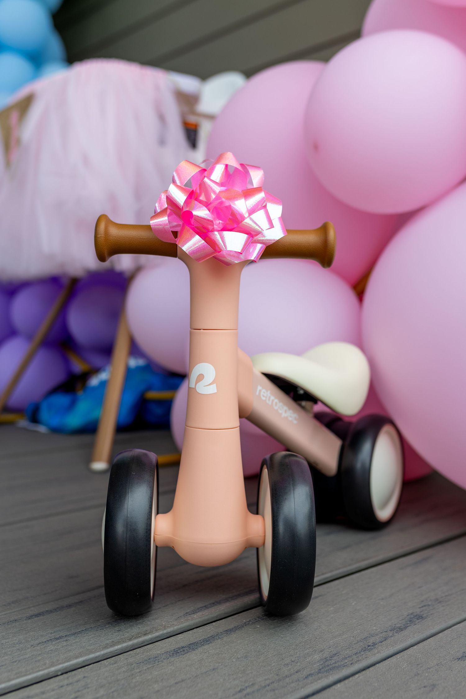 Pink decorated balance bike with bow against pastel balloon backdrop.
