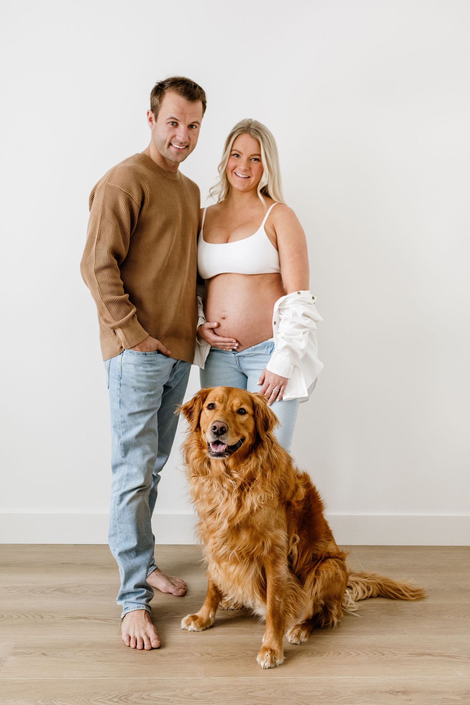 A couple sits with their Golden Retriever dog on a wooden floor during a maternity photo session.