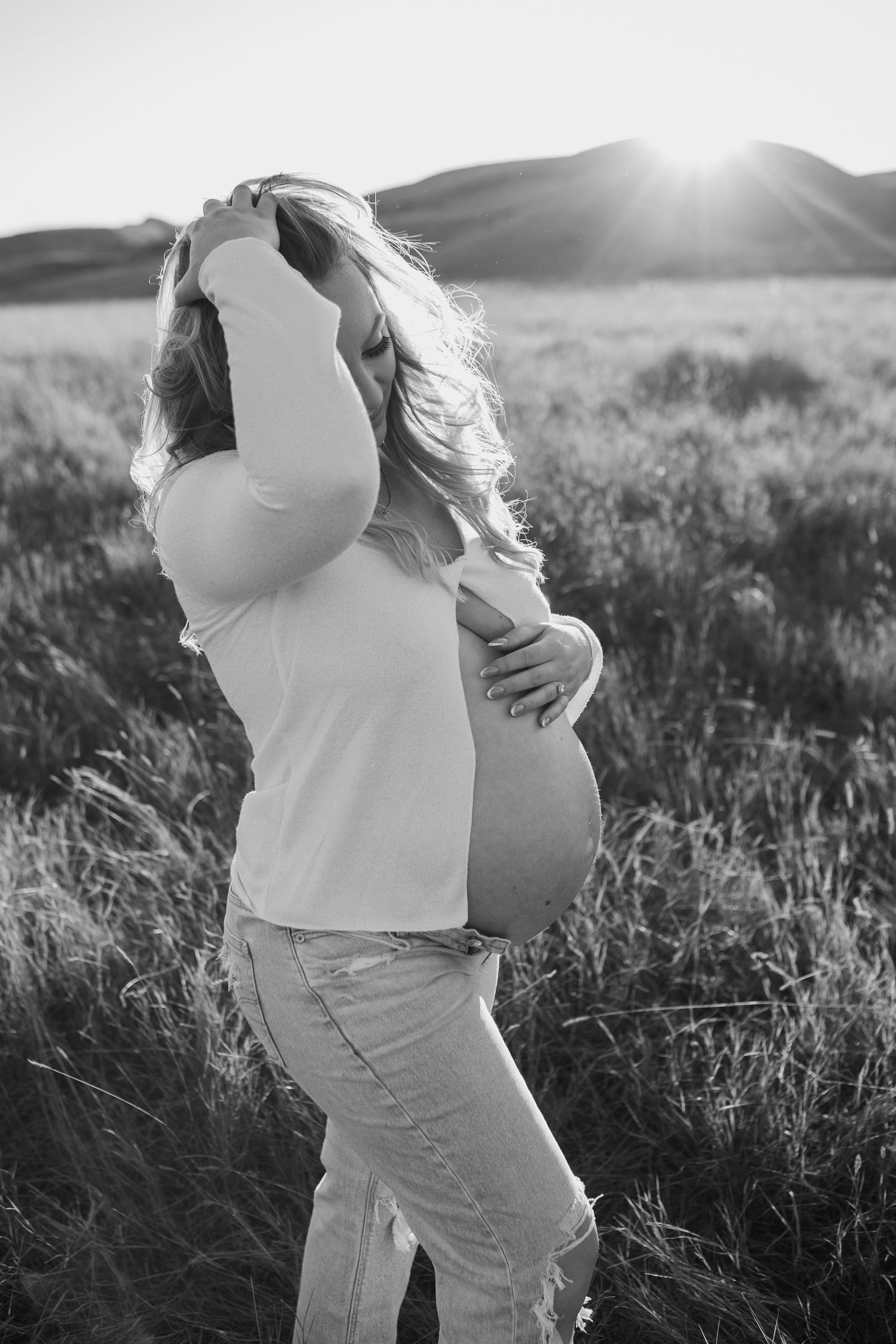 Silhouette of expectant mother standing in a sunlit field of tall grass at sunset.