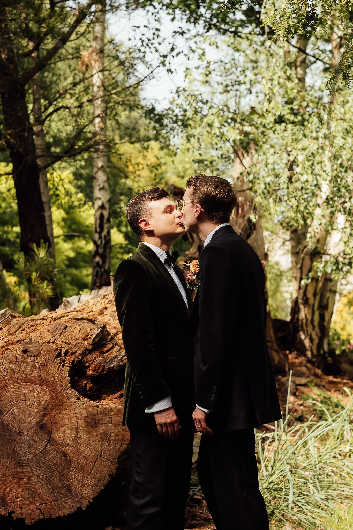 Two grooms in black suits share a kiss outdoors near a large log in a lush forest wedding setting.