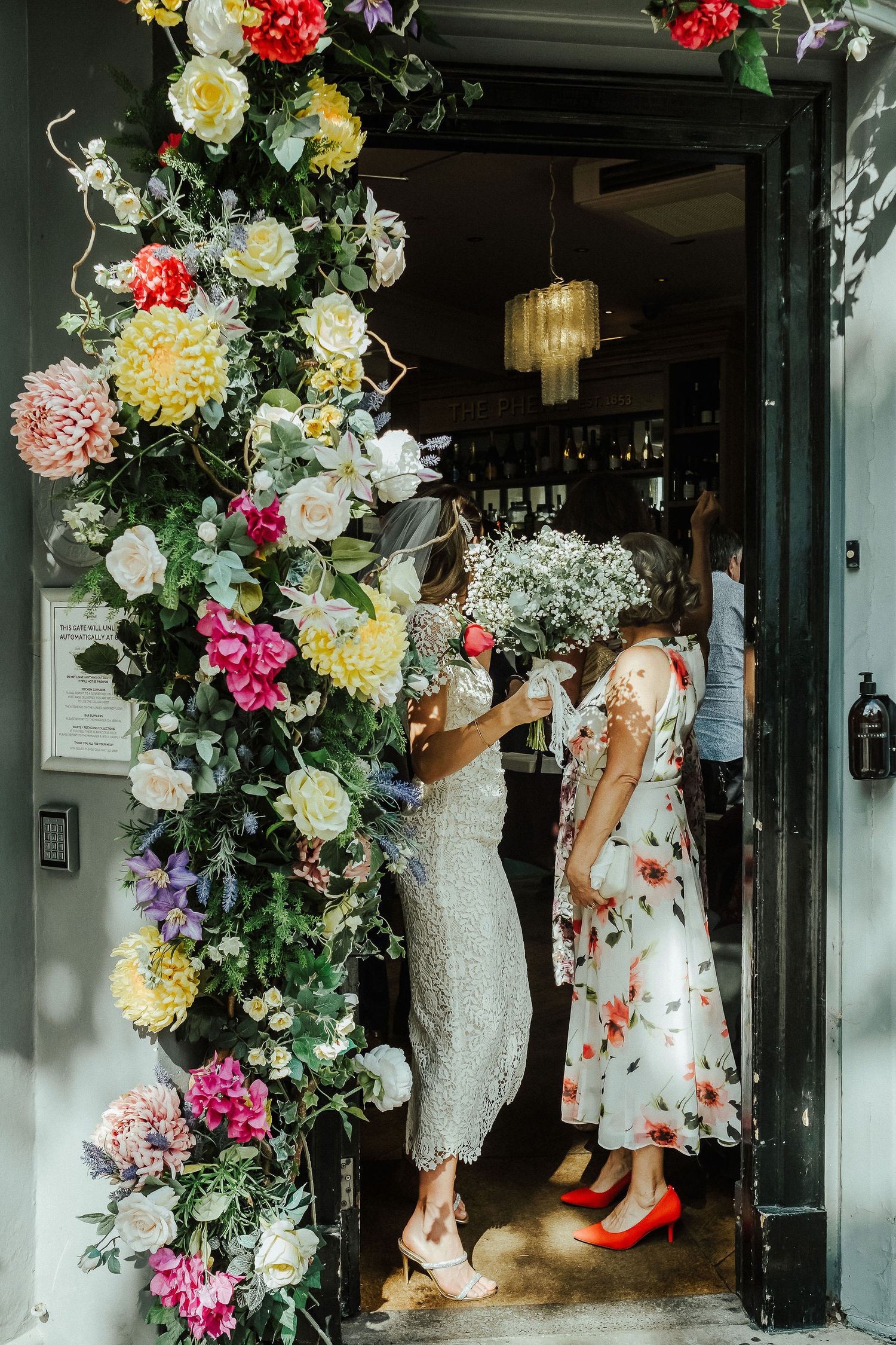 Bridesmaids in floral and white dresses gather in a hallway decorated with colorful flower arrangements and greenery.