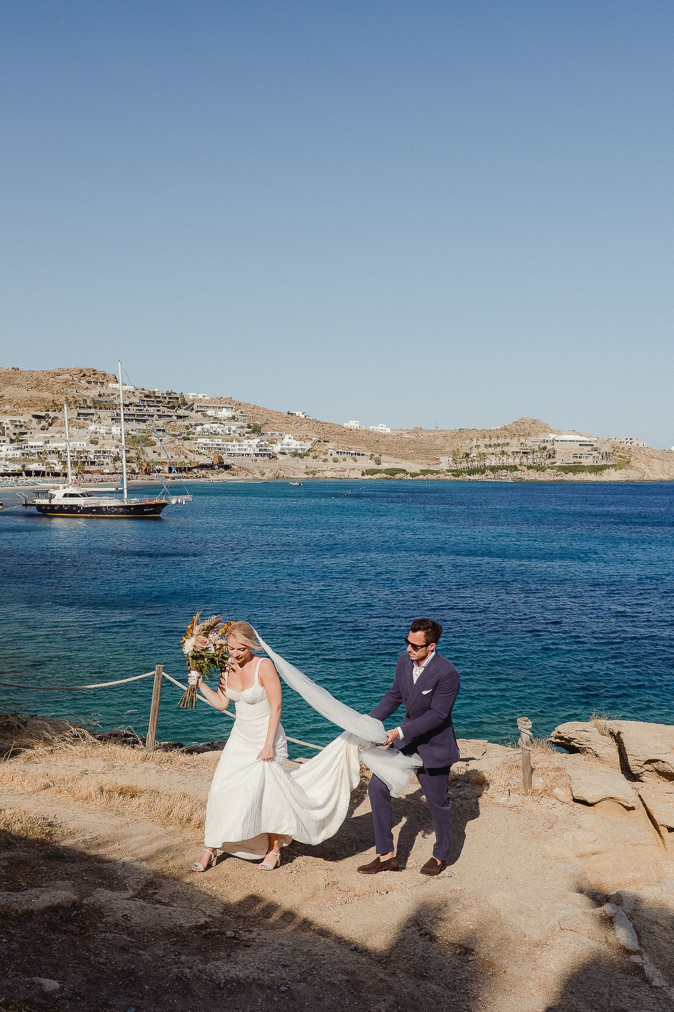 Couple sits together on rocky shoreline overlooking blue Mediterranean waters and coastal hills on sunny day.