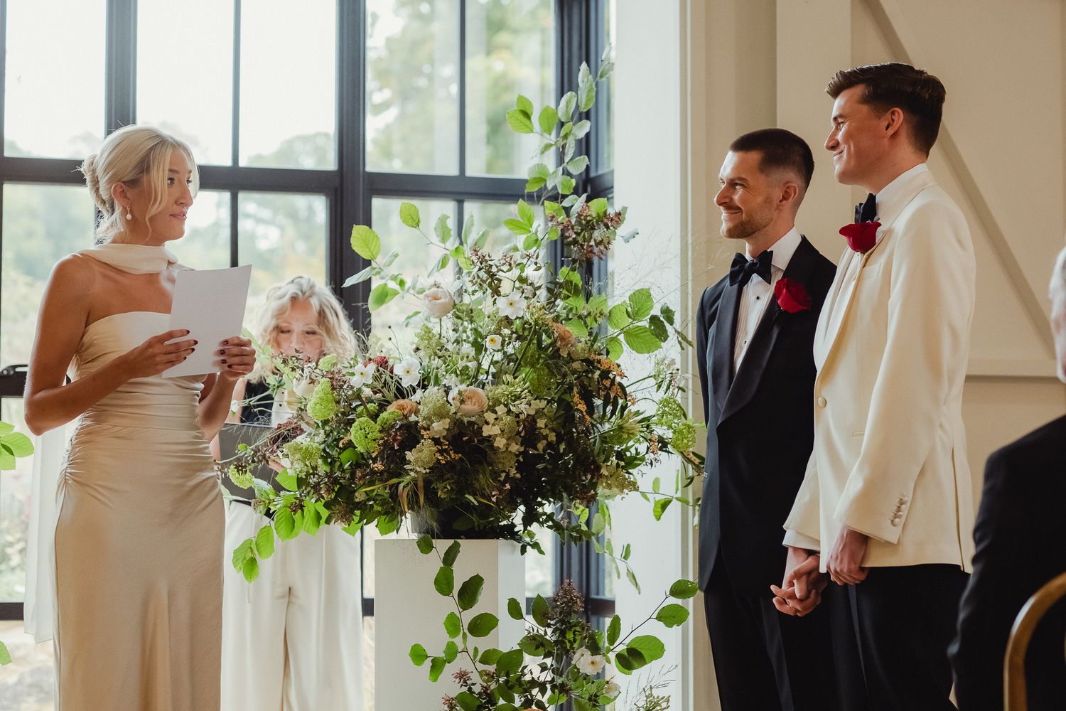 A wedding party stands in a bright indoor venue with large windows and floral decorations.