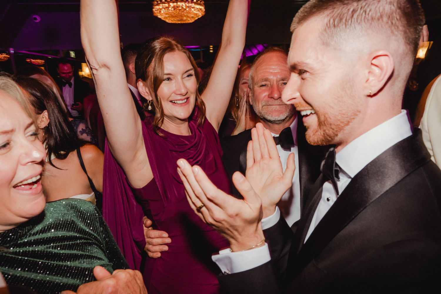 Wedding guests dancing and celebrating enthusiastically on a lively dance floor with purple lighting.
