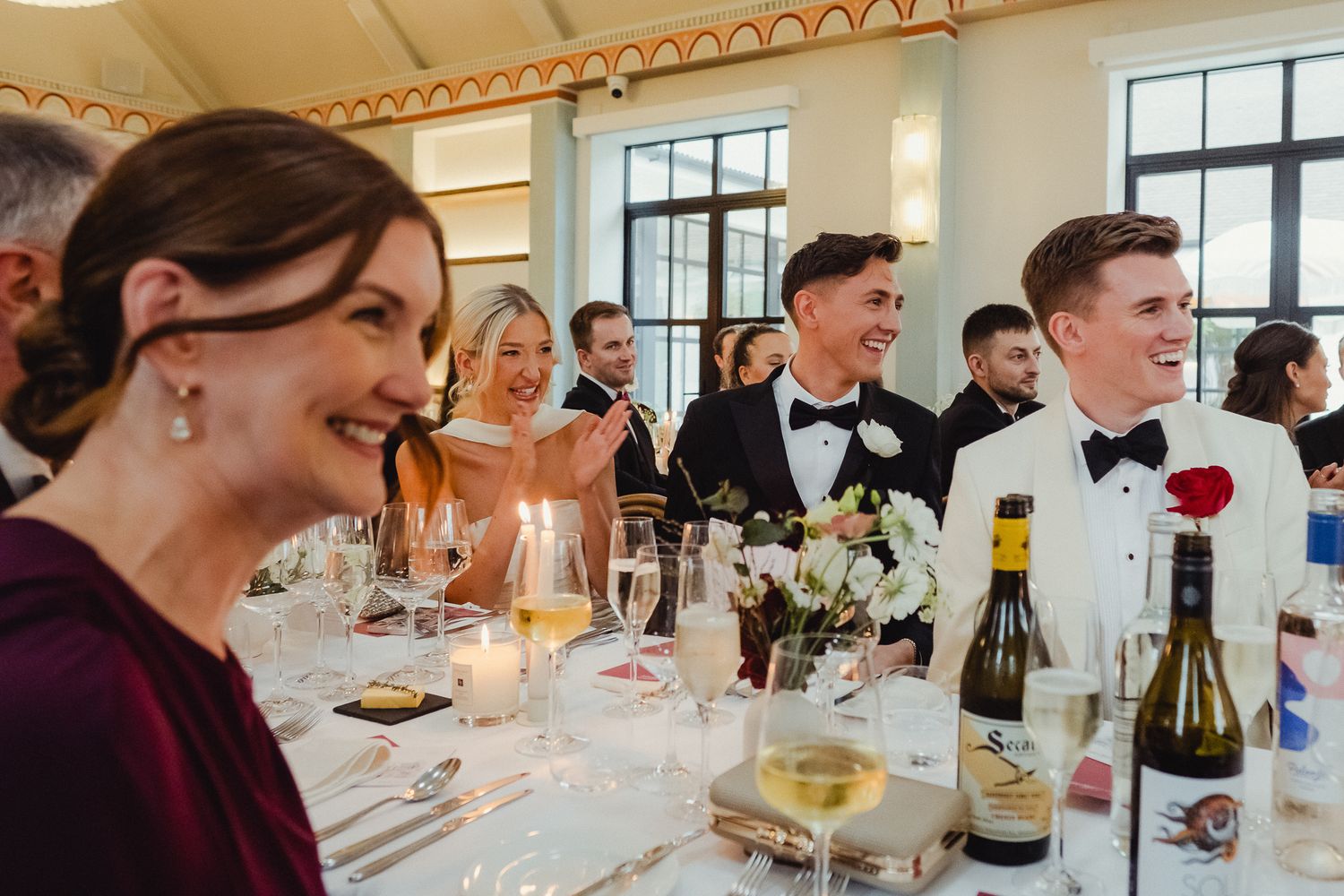 Wedding guests enjoy a candlelit dinner reception with wine bottles and elegant table settings at a formal venue.