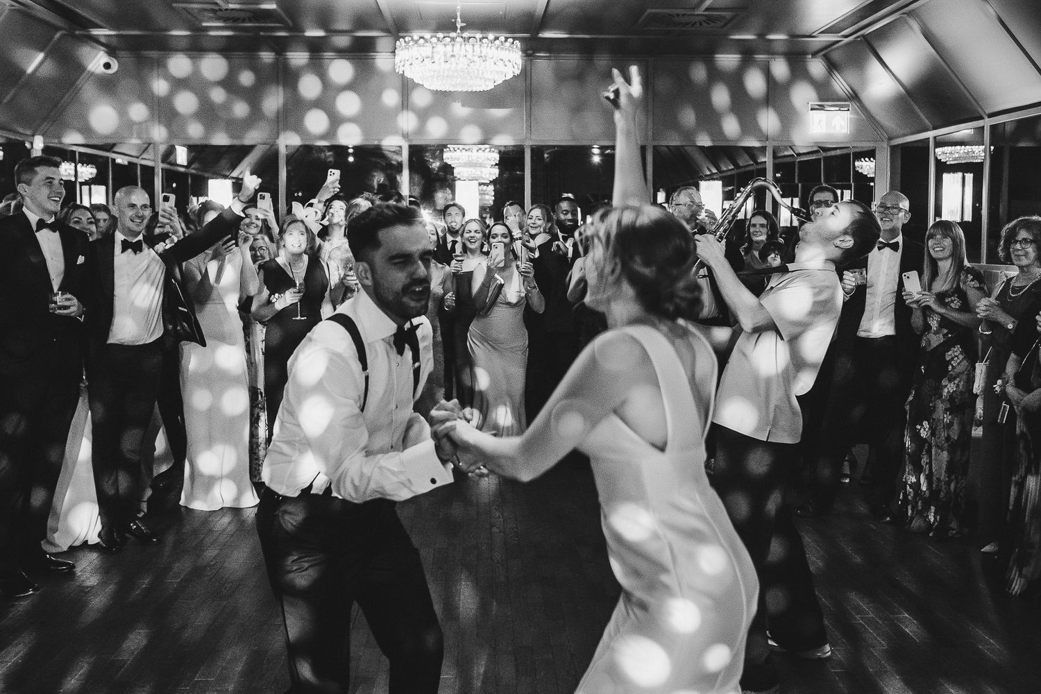 Wedding guests dance together on a dark dance floor illuminated by spotlights during an evening reception celebration.