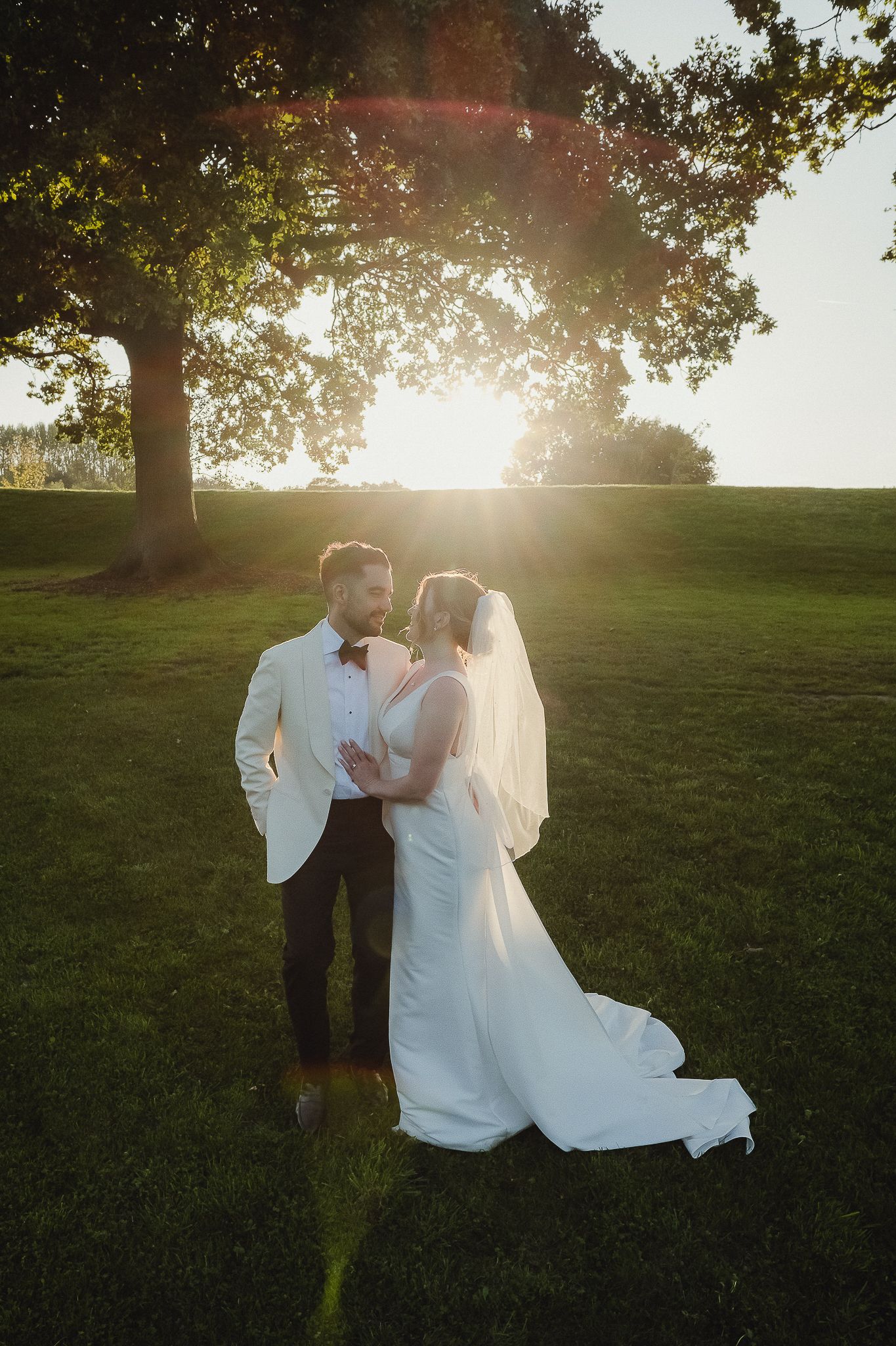 A couple in wedding attire embraces on a grassy hill during golden hour sunset.